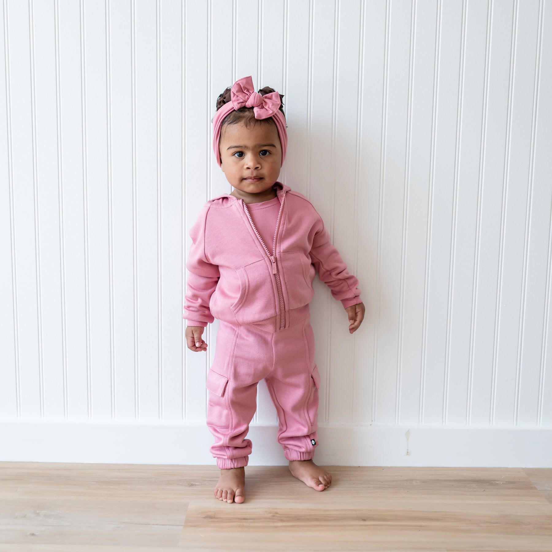 Young toddler standing in front of a white paneled wall wearing the Fleece Zip Up Hoodie in Apple Blossom and matching fleece cargo pants and Knotted bow