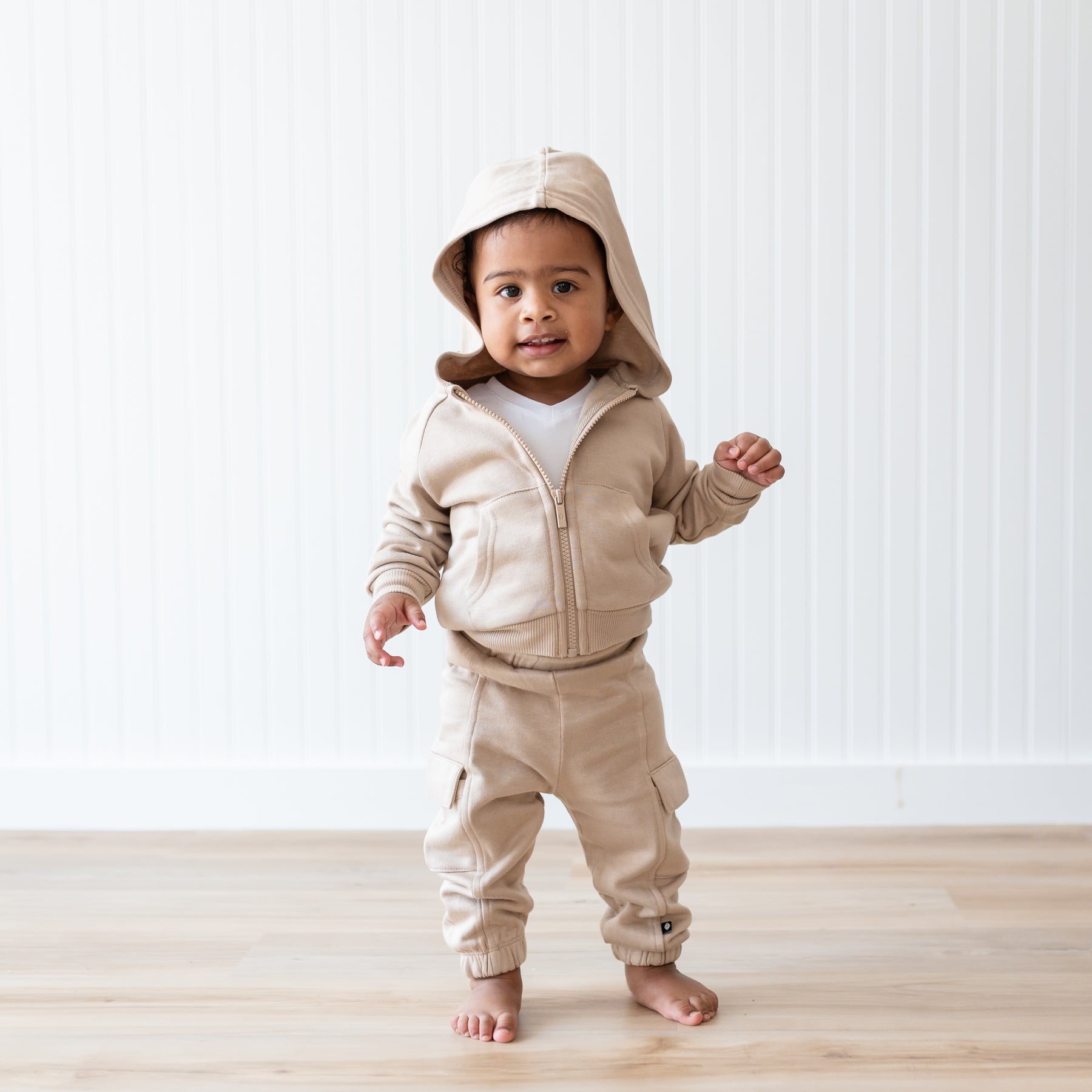 Young toddler wearing the Fleece Zip Up Hoodie in Almond with the hood up and matching Fleece Cargo pants standing in front of a white paneled wall