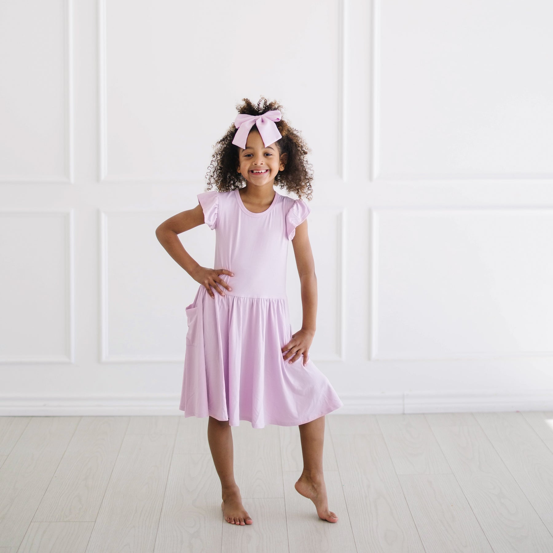 Young girl posing in front of a white wall wearing the Pocket Dress in Thistle with matching large alligator bow clip
