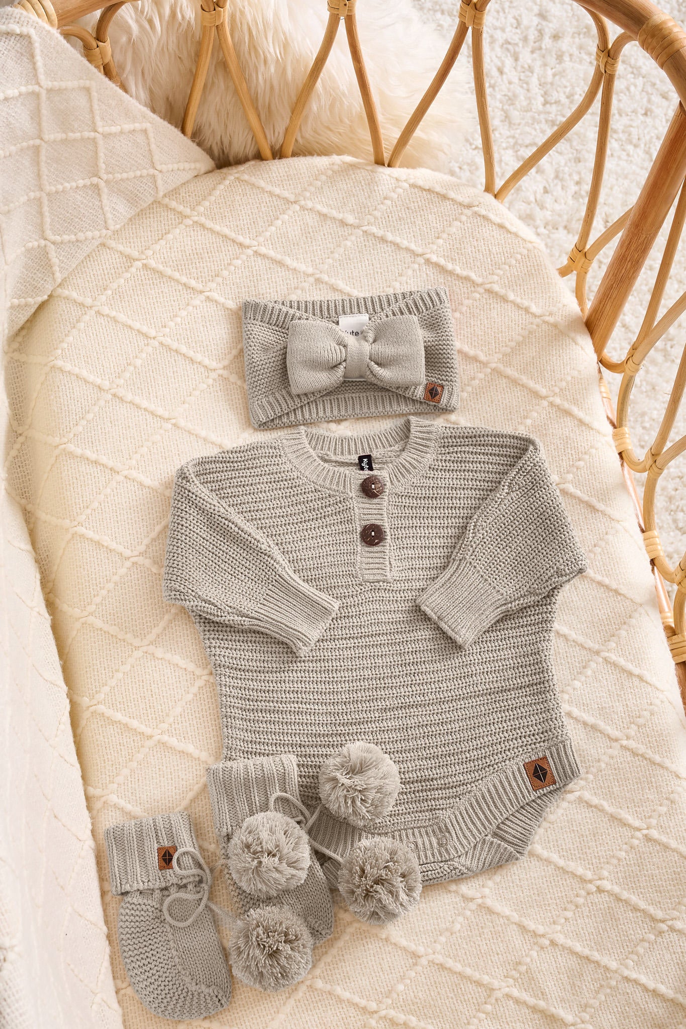 A baby knit outfit set in a light gray tan, including a long-sleeve henley bodysuit with three brown buttons, a matching bow headband, and a pair of booties with pom-poms, all arranged on a cream quilted blanket inside a wicker basket.