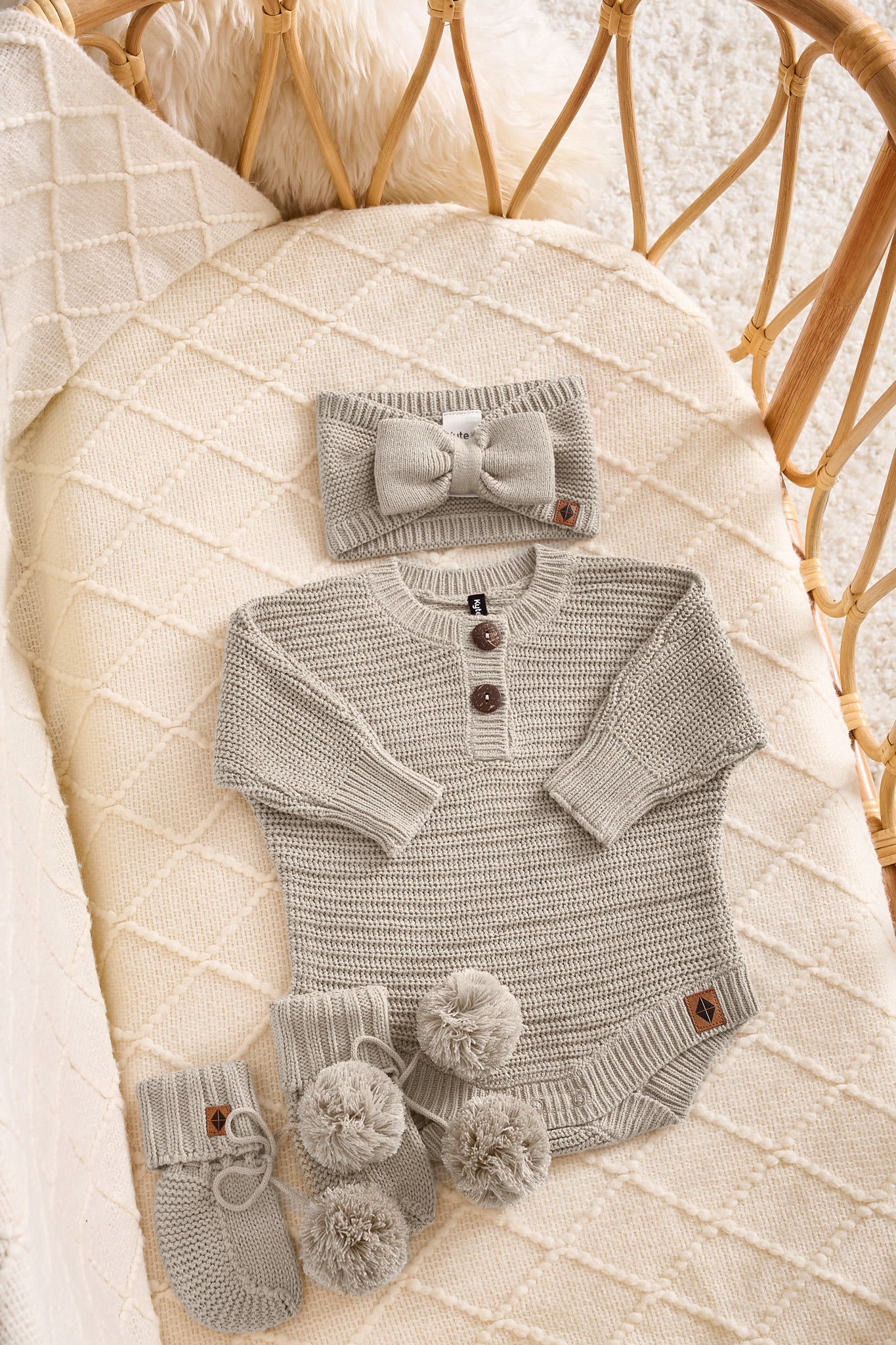A baby knit outfit set in a light gray tan, including a long-sleeve henley bodysuit with three brown buttons, a matching bow headband, and a pair of booties with pom-poms, all arranged on a cream quilted blanket inside a wicker basket.