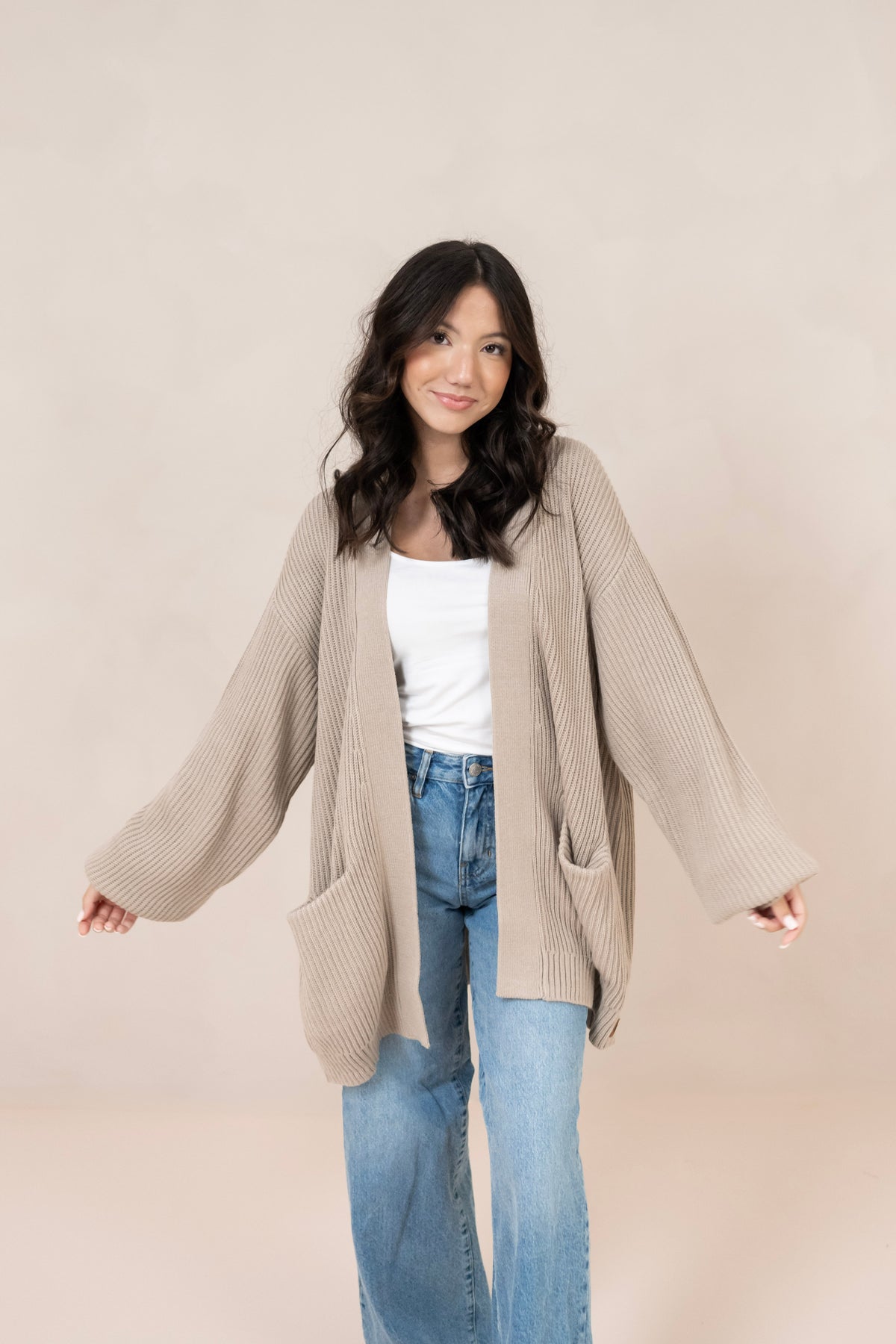 Woman wearing a beige cardigan over a white top and blue jeans on a plain background