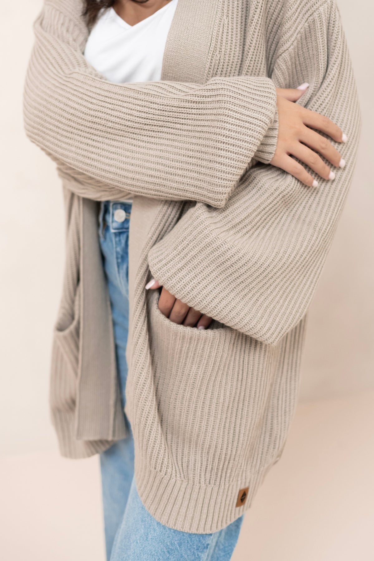 Person wearing a beige cardigan over a white shirt and blue jeans on a plain background
