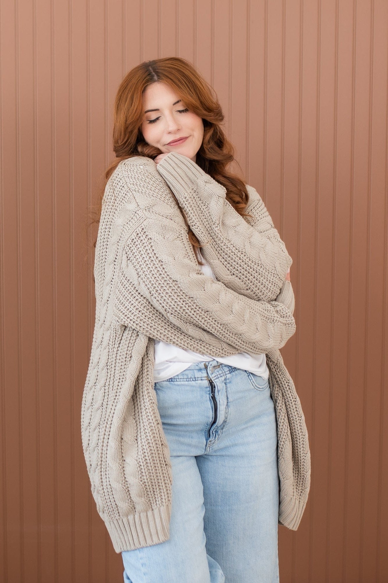 Female model hugging herself wearing the Cable Knit Adult Cardigan in Almond paired with light wash jeans