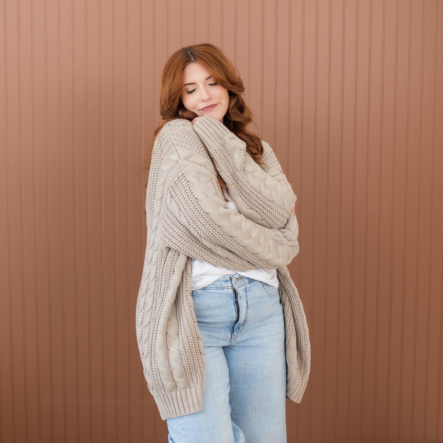 Female model hugging herself wearing the Cable Knit Adult Cardigan in Almond paired with light wash jeans