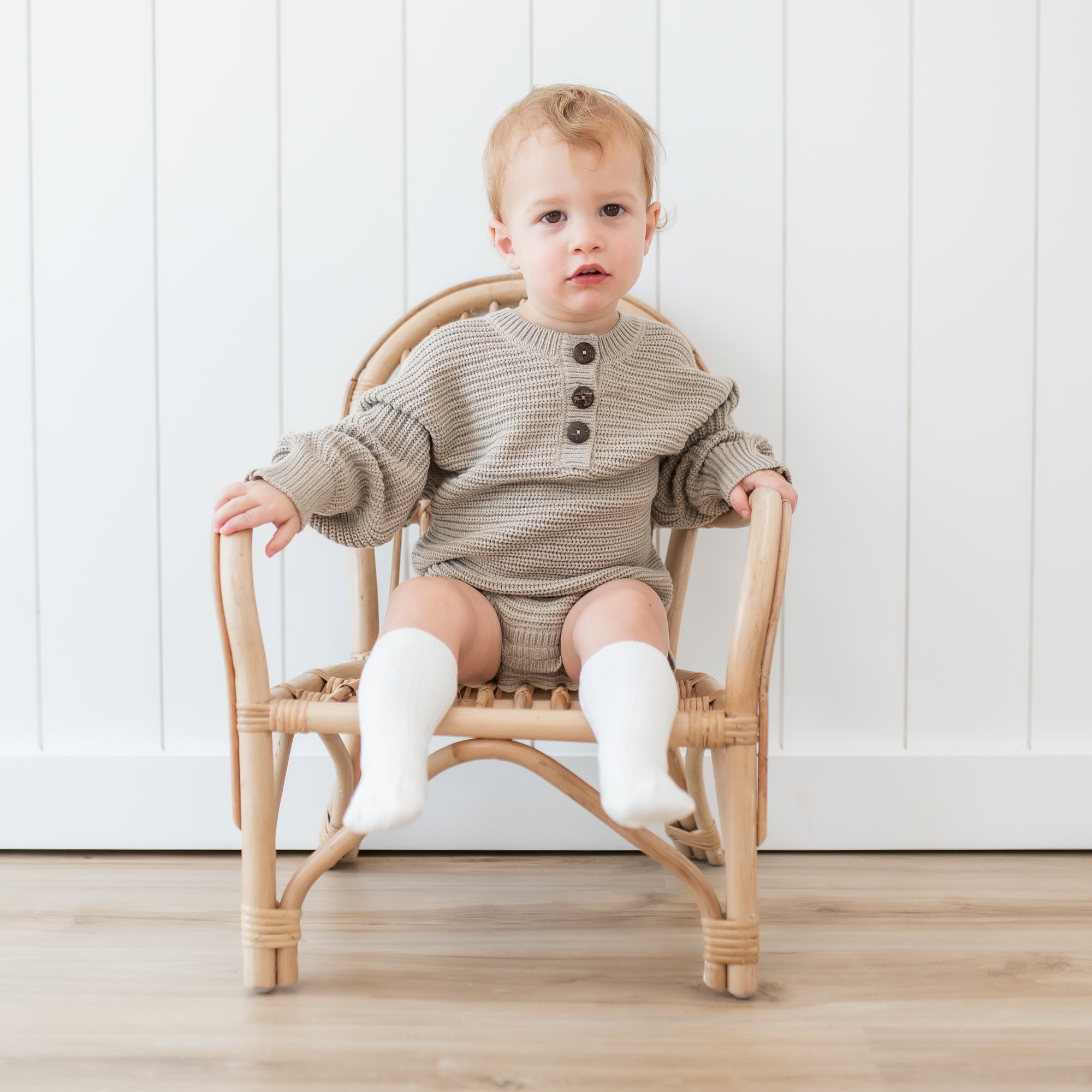 Toddler sitting in a wicker chair wearing the Chunky Knit Romper in Almond and Cloud Knee high socks