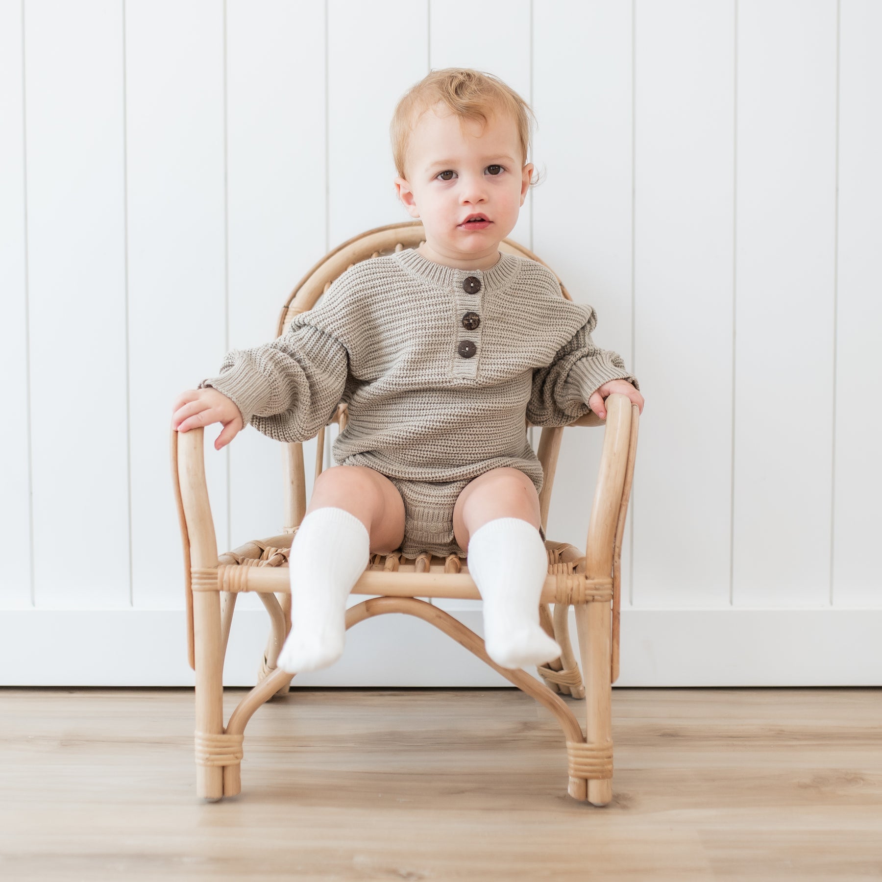 Toddler sitting in a wicker chair wearing the Chunky Knit Romper in Almond and Cloud Knee high socks