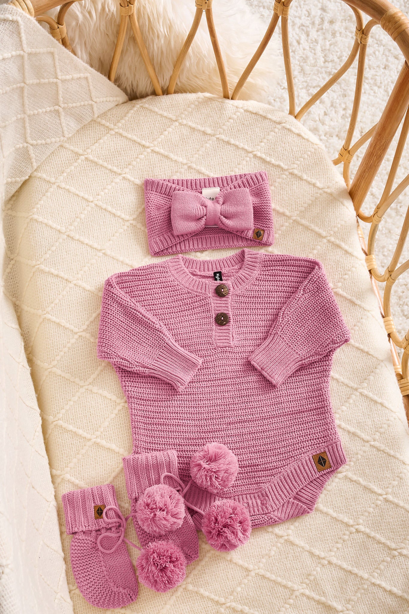 A pink knit baby outfit set arranged on a cream quilted blanket in a rattan bassinet. The set includes a long-sleeve henley bodysuit with three buttons, a matching bow headband, and a pair of booties with pom-poms.