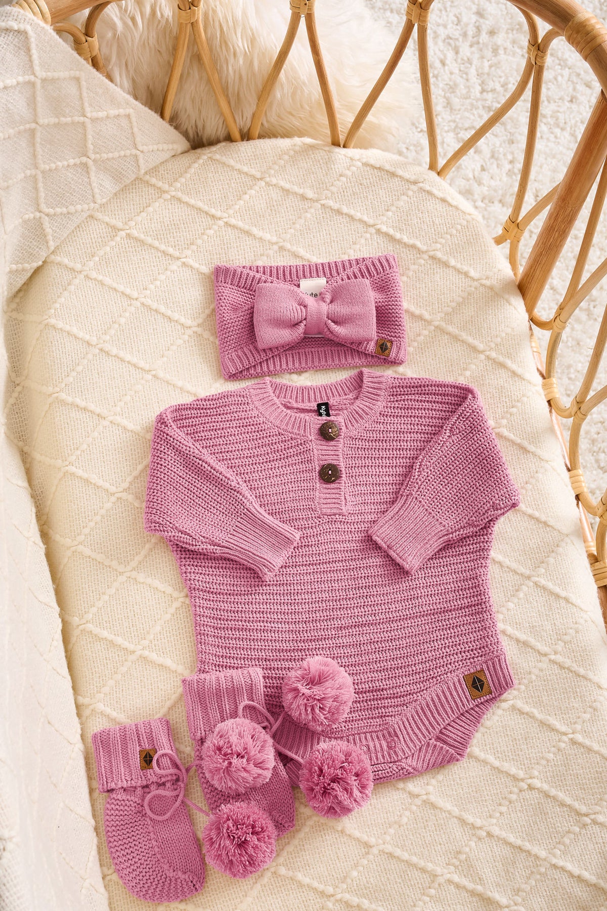A pink knit baby outfit set arranged on a cream quilted blanket in a rattan bassinet. The set includes a long-sleeve henley bodysuit with three buttons, a matching bow headband, and a pair of booties with pom-poms.