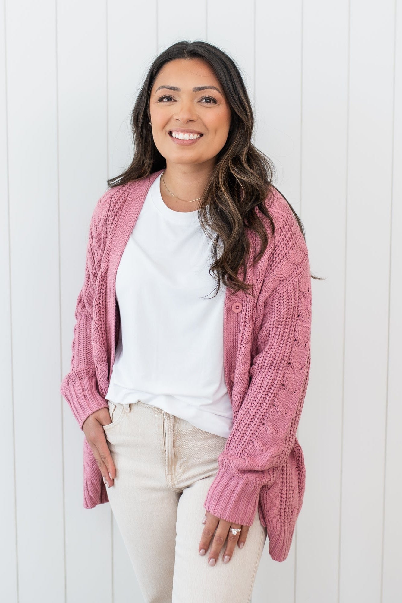 Female wearing the Cable Knit Adult Cardigan in Apple Blossom paired with a white shirt and beige colored pants