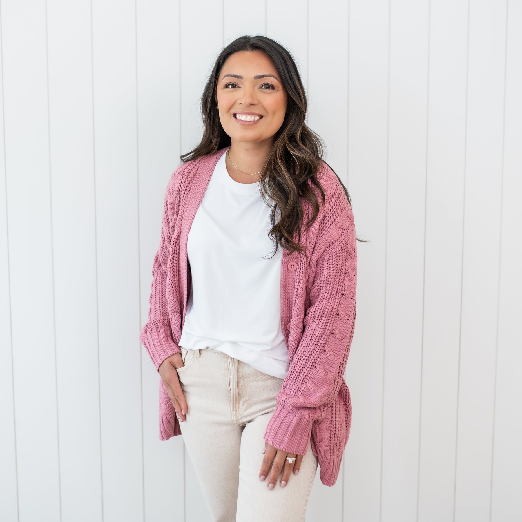 Female wearing the Cable Knit Adult Cardigan in Apple Blossom paired with a white shirt and beige colored pants