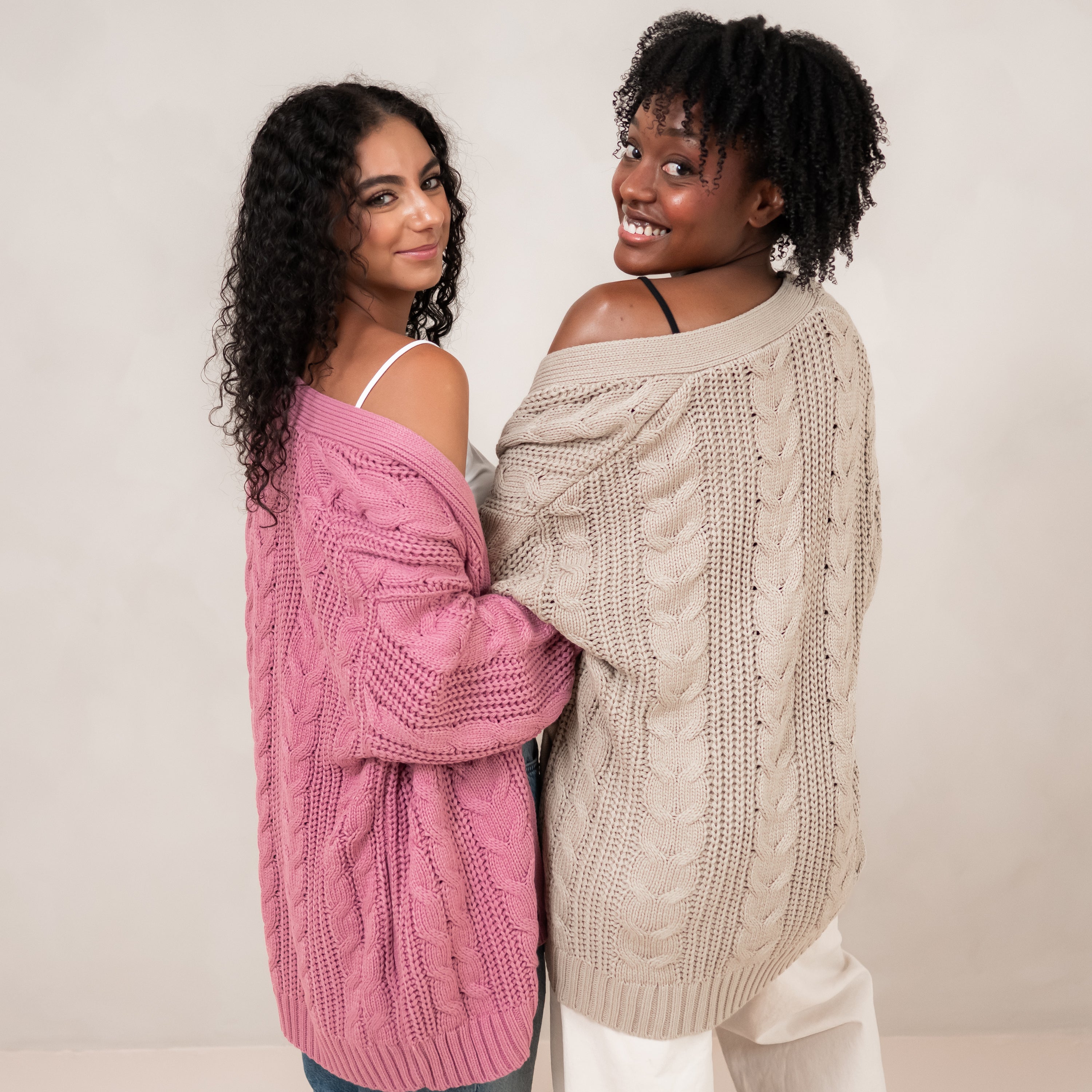 Two models standing side by side with their backs to the camera wearing the Cable Knit Adult Cardigan in Almond and Apple Blossom
