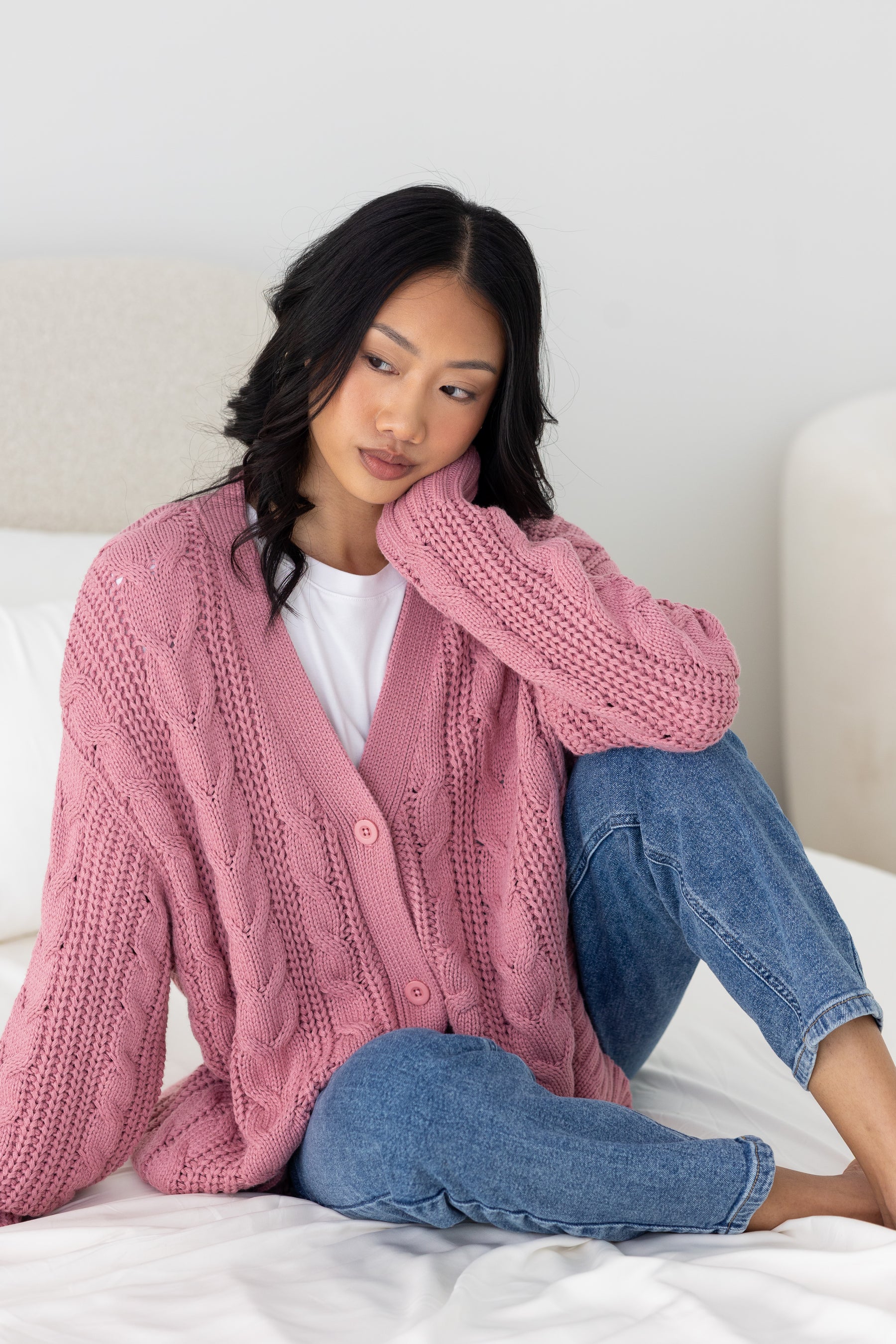 Woman wearing a pink knitted cardigan and blue jeans sitting on a bed.