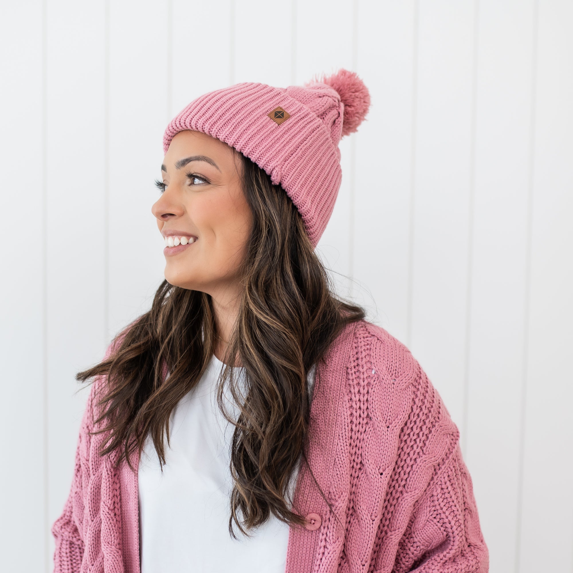 Smiling female model wearing the Cable Knit Adult Pom Beanie in Apple Blossom with matching cable knit cardigan