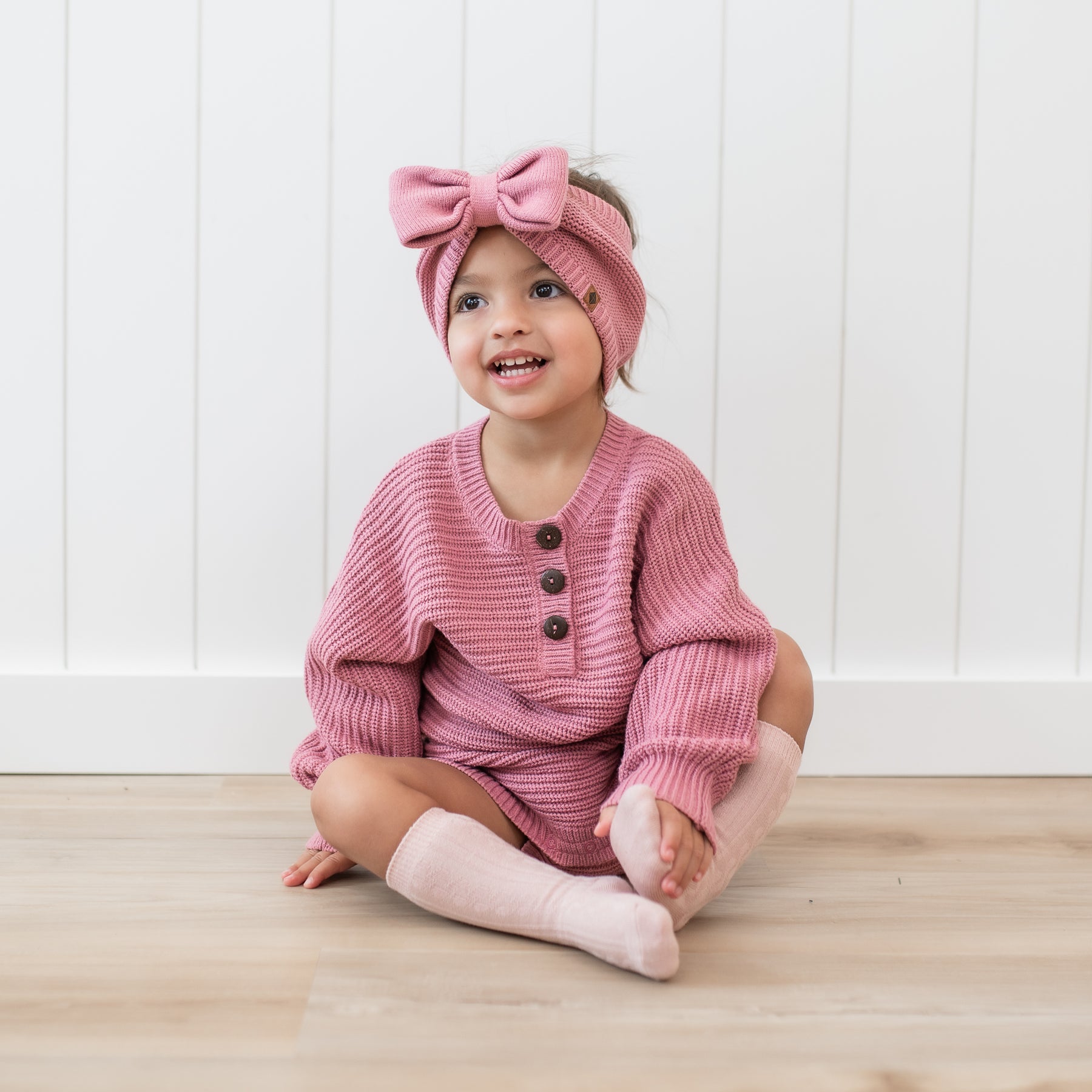 Toddler sitting wearing the Chunky Knit Bow Headband in Apple Blossom paired with a matching chunky knit romper
