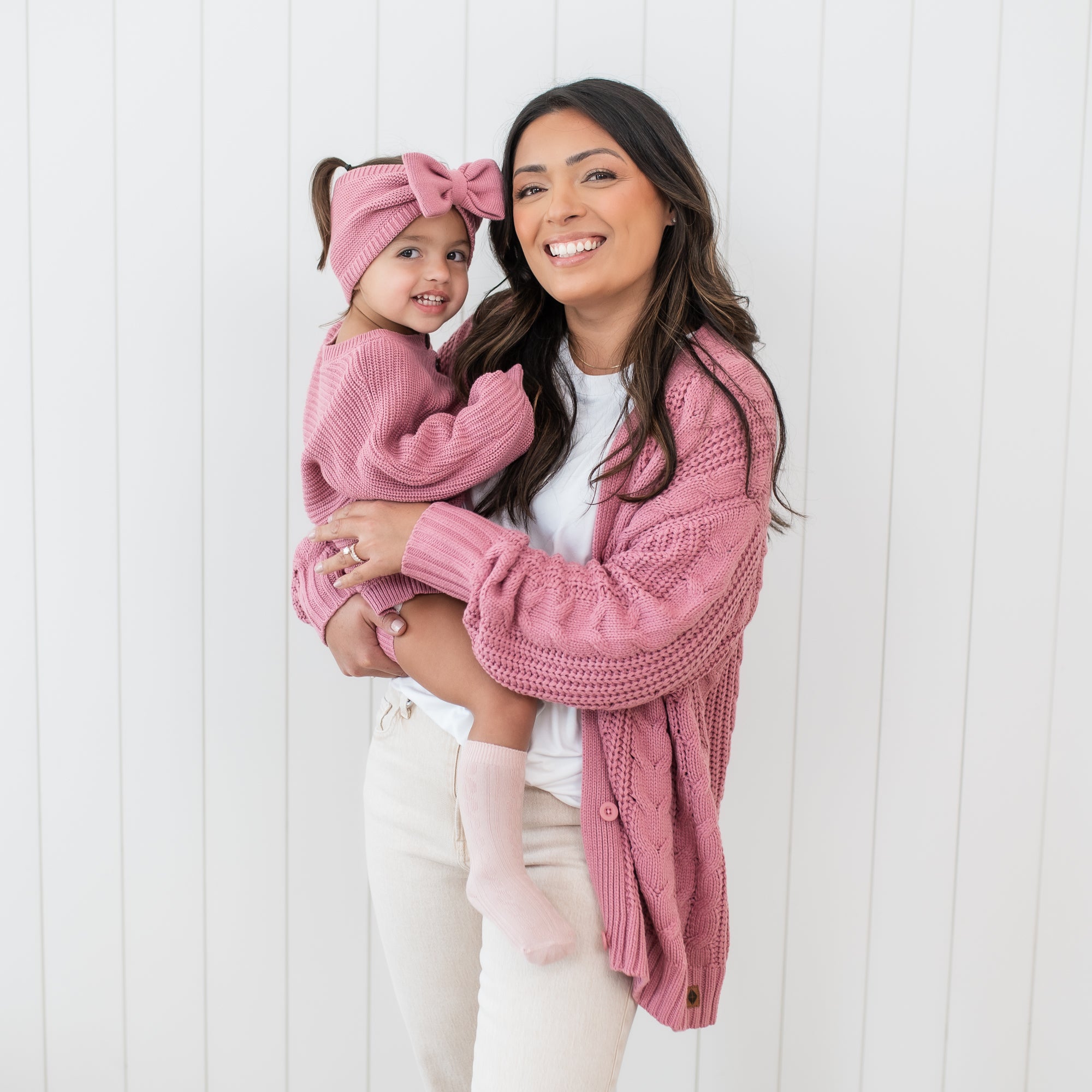 Smiling mother wearing the Cable Knit Cardigan in Apple blossom holding her daughter who is wearing the Chunky Knit Romper in Apple Blossom and matching chunky knit bow headband