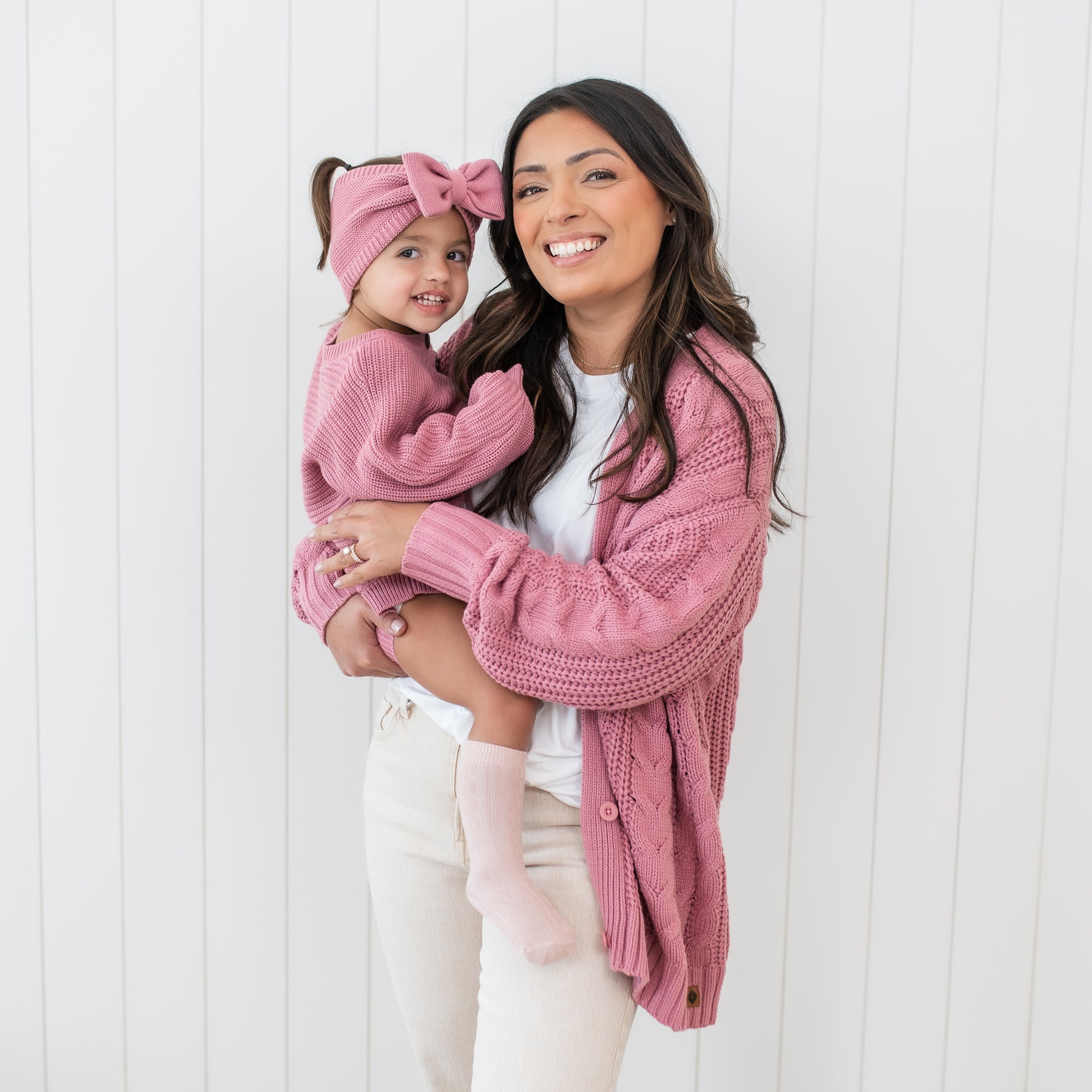 Smiling mother wearing the Cable Knit Cardigan in Apple blossom holding her daughter who is wearing the Chunky Knit Romper in Apple Blossom and matching chunky knit bow headband