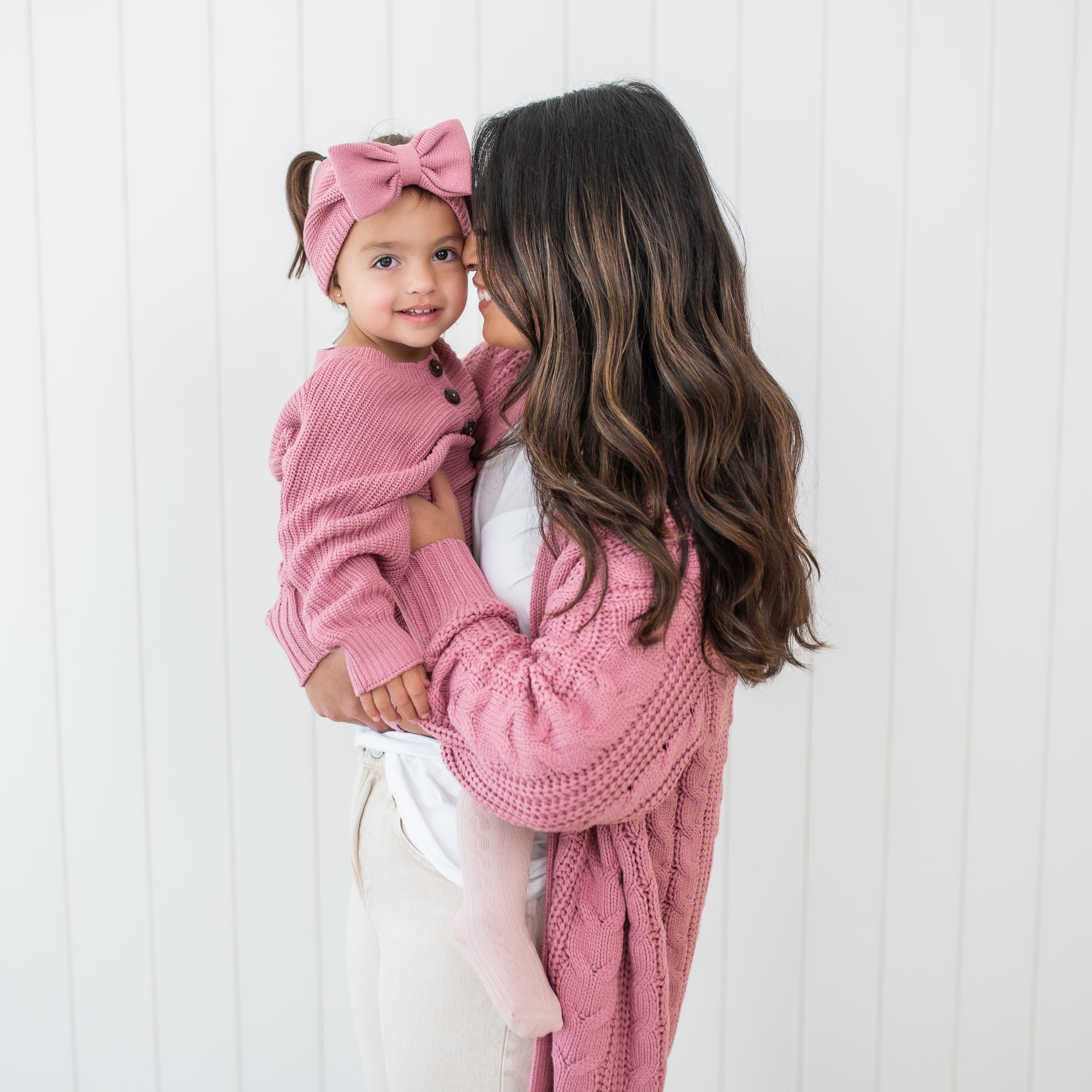 Mother, wearing the Cable Knit Cardigan in Apple Blossom, holding daughter who is wearing the Chunky Knit Bow Headband in Apple Blossom and matching Chunky Knit romper