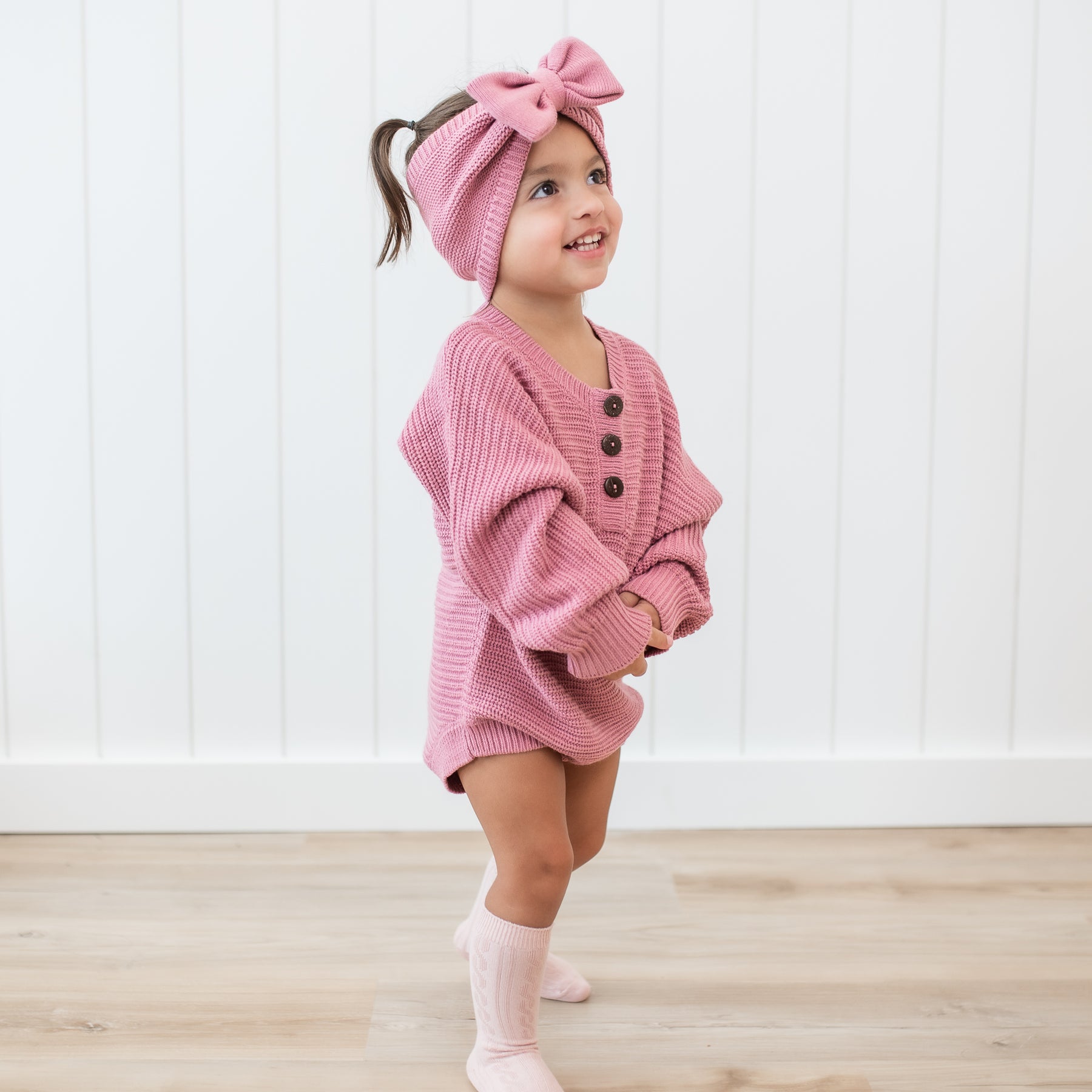 Smiling toddler wearing the Chunky Knit Romper in Apple Blossom with matching chunky knit bow headband