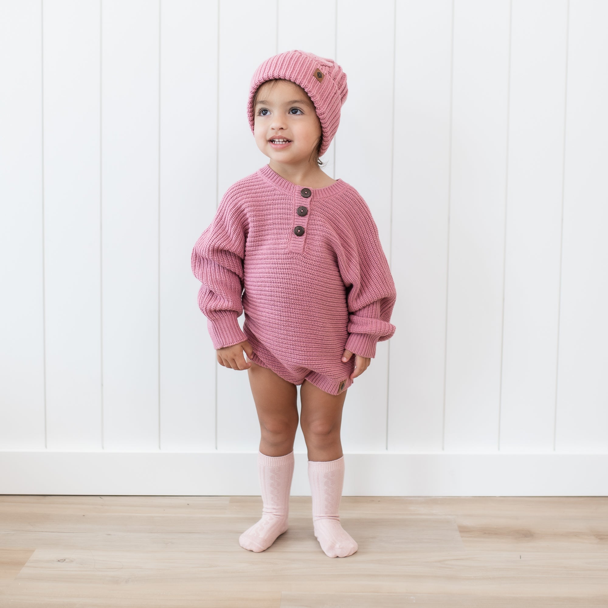 Toddler wearing the Chunky Knit Romper in Apple Blossom with matching cable knit pom beanie standing in front of a white paneled wall