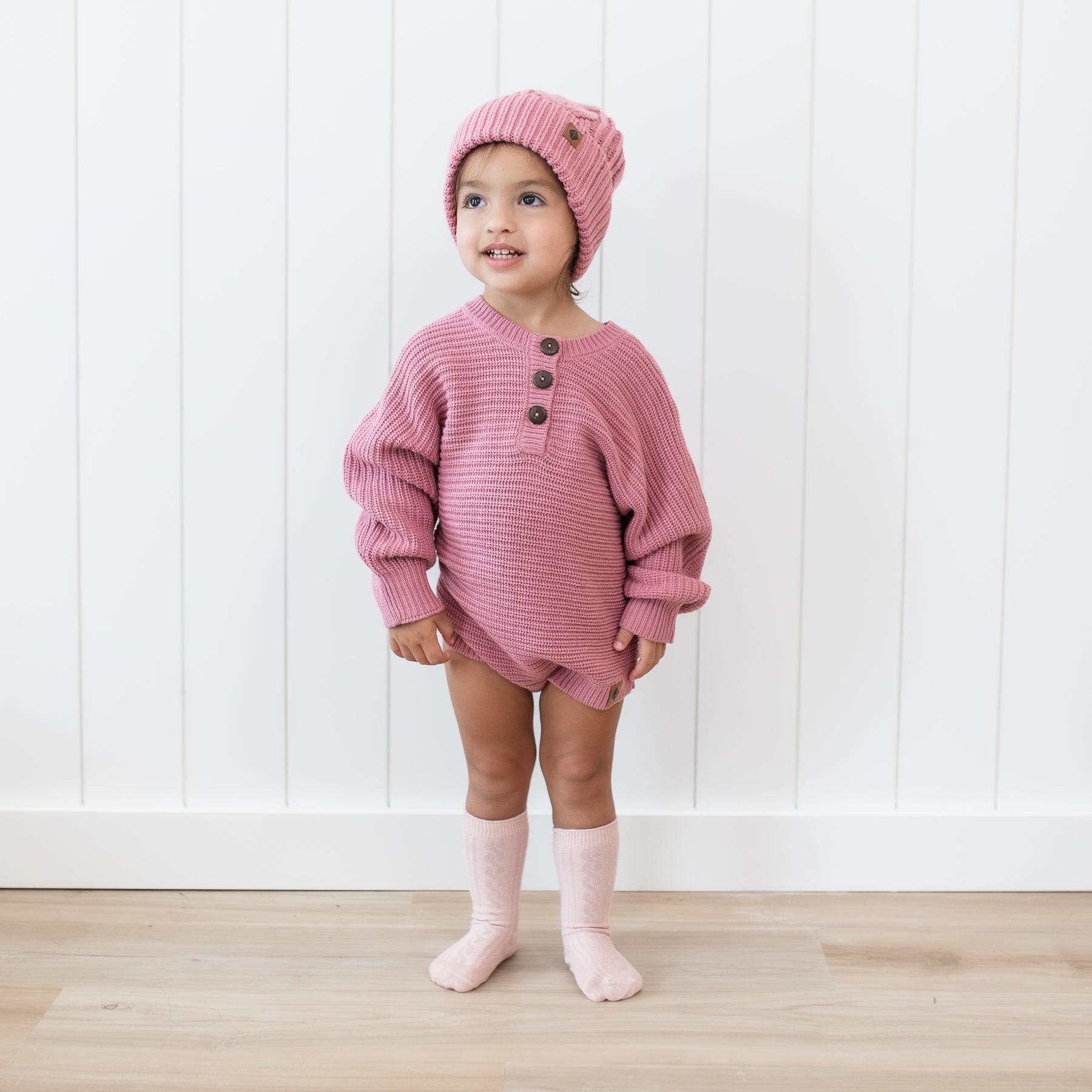 Toddler wearing the Chunky Knit Romper in Apple Blossom with matching cable knit pom beanie standing in front of a white paneled wall