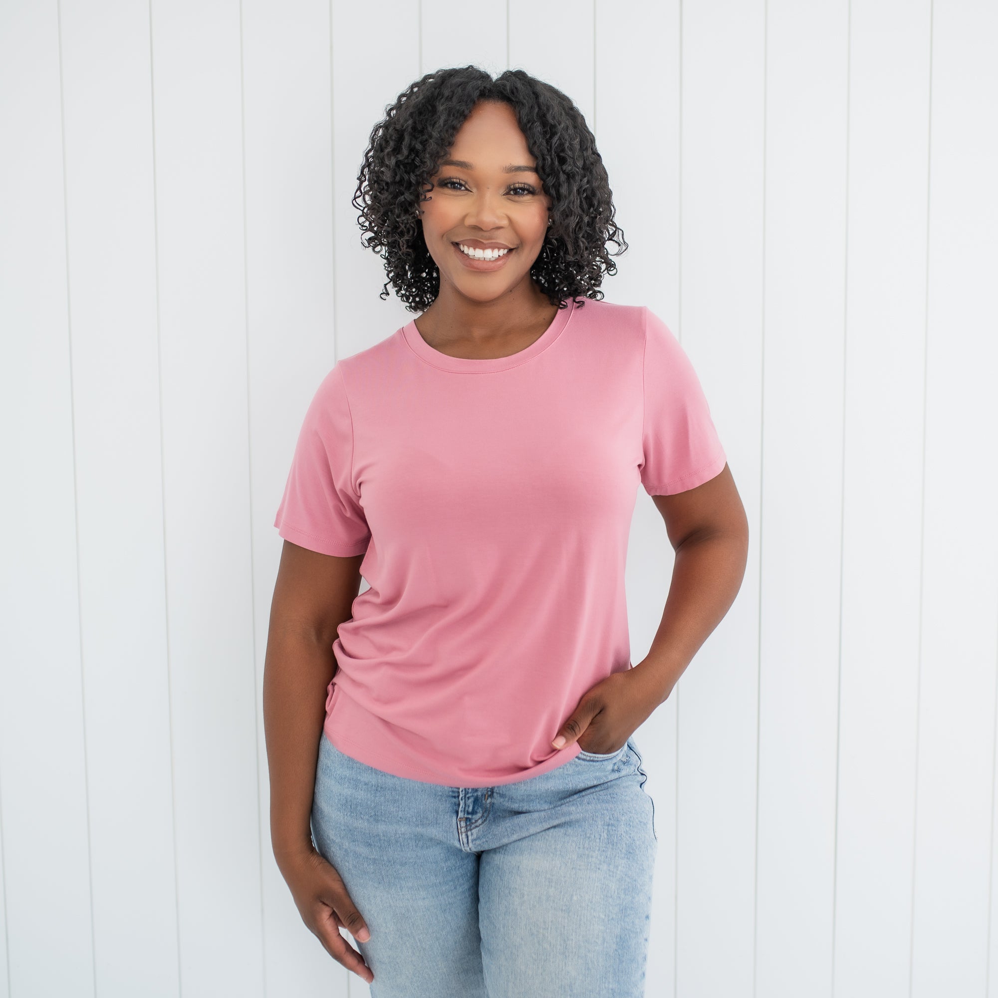 Female standing wearing the Women's Crew Neck Tee in Apple Blossom with one hand in her jean pocket in front of a white paneled wall