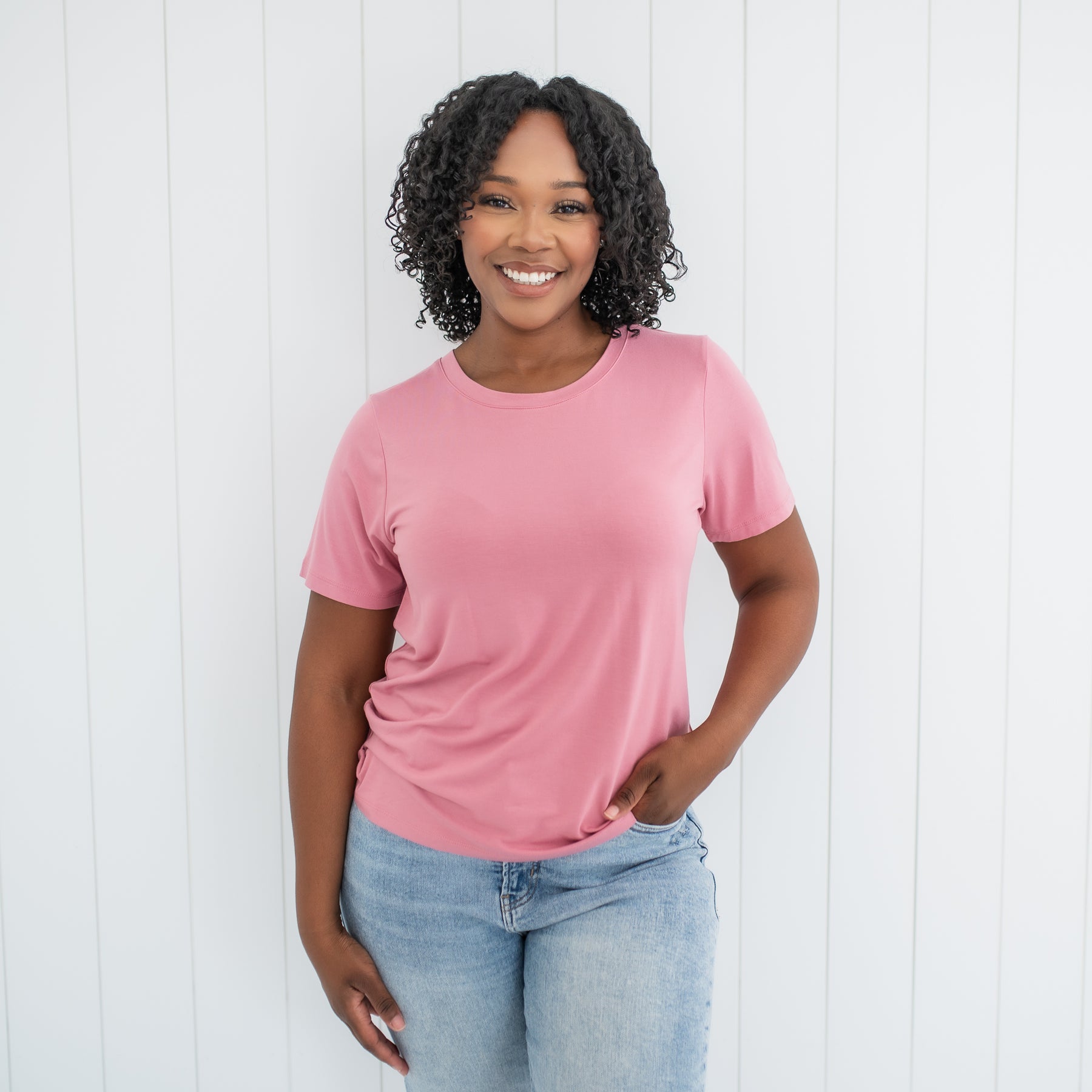 Female standing wearing the Women's Crew Neck Tee in Apple Blossom with one hand in her jean pocket in front of a white paneled wall