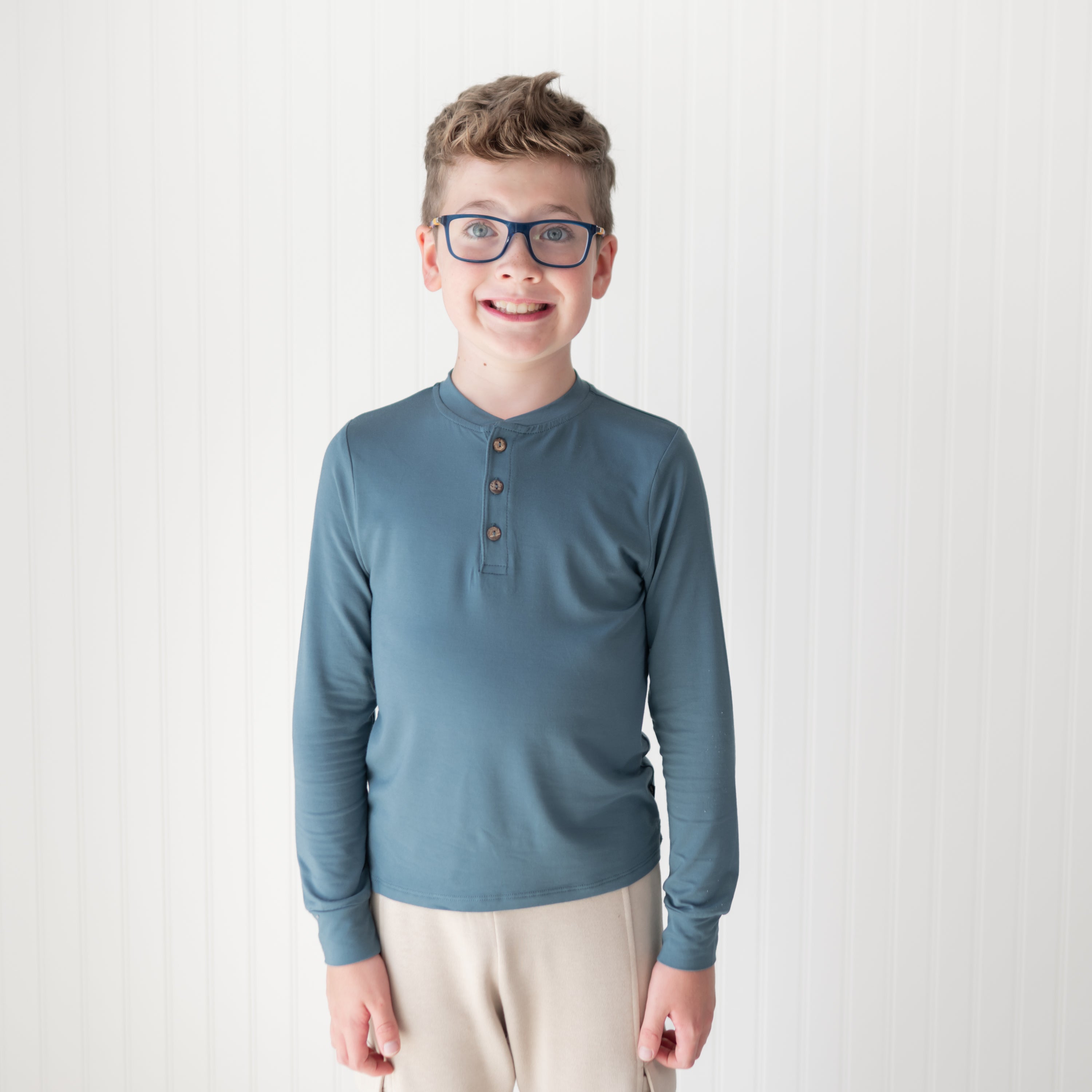 Young boy wearing the Long Sleeve Toddler Henley Tee in Atlantic standing in front of a white paneled wall