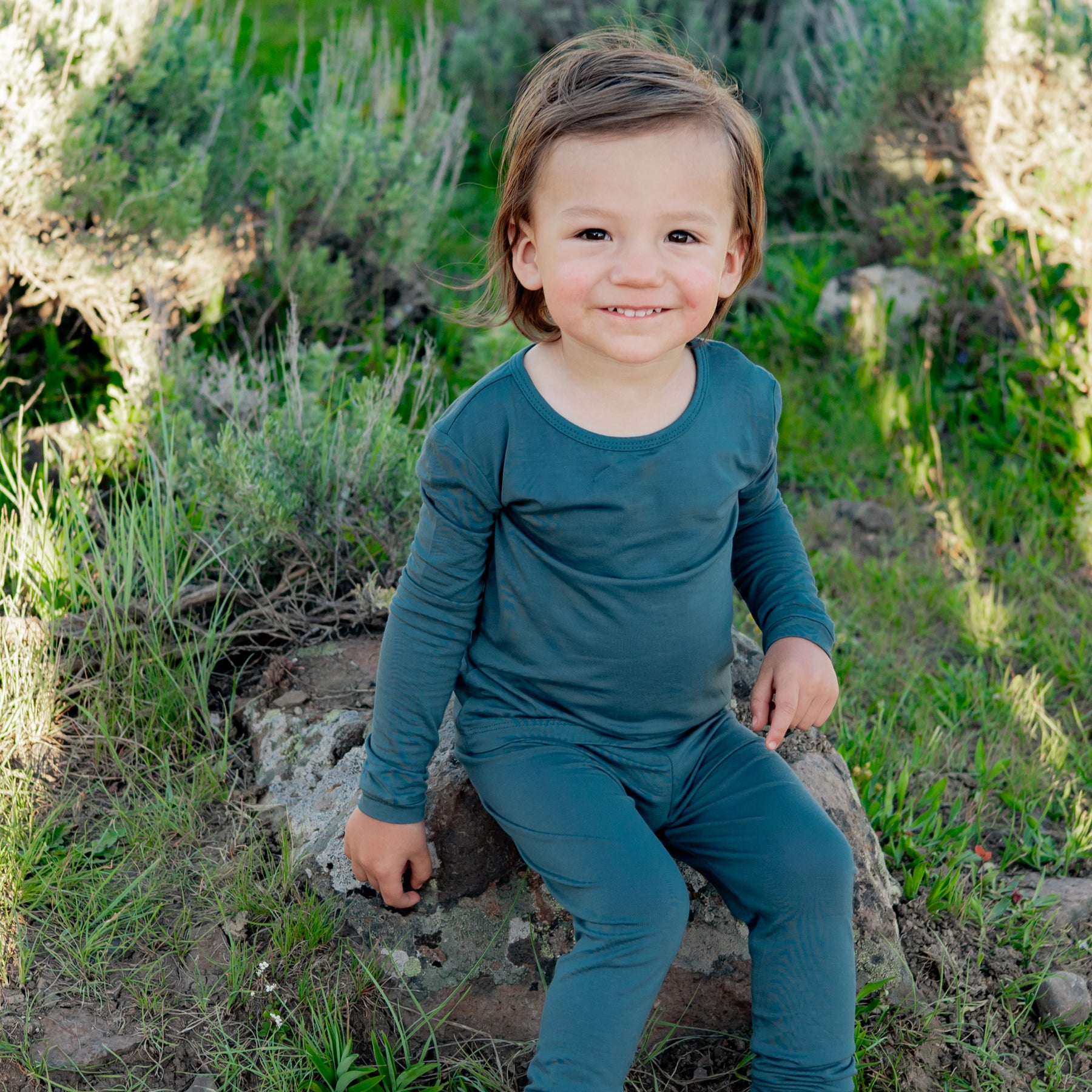 Smiling toddler sitting on a rock outside wearing the Long Sleeve Pajamas in Atlantic