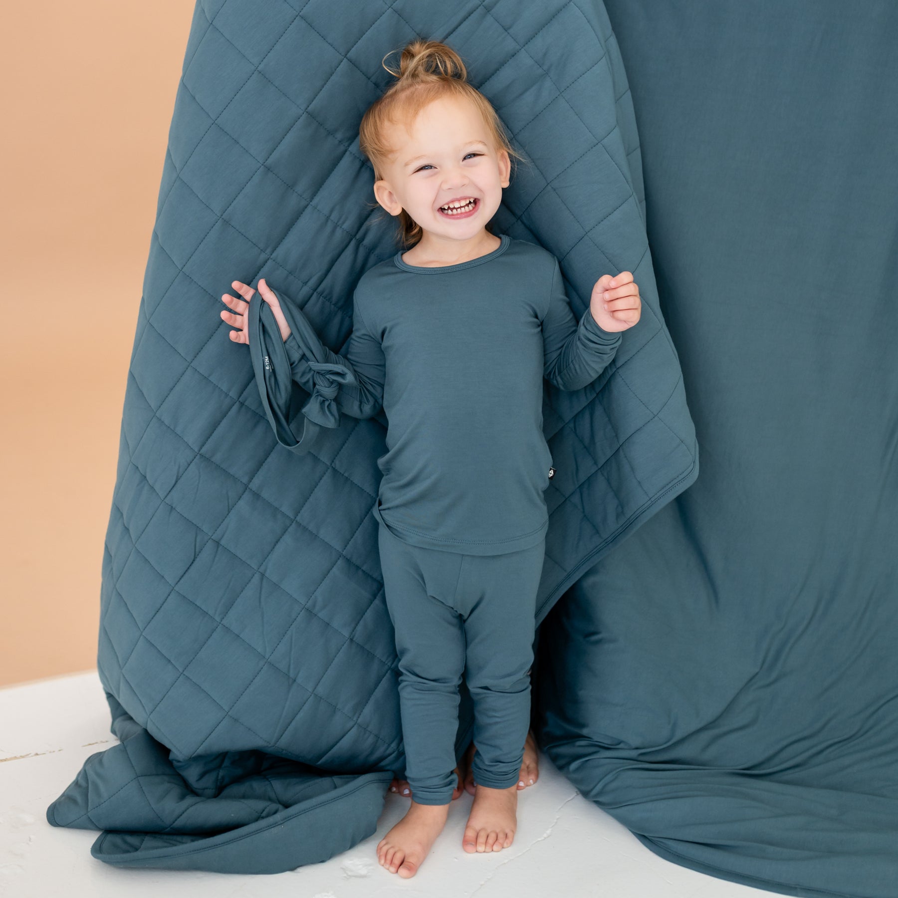 Smiling toddler standing in front of an Atlantic blanket holding a knotted bow headband wearing the Long Sleeve Pajamas in Atlantic