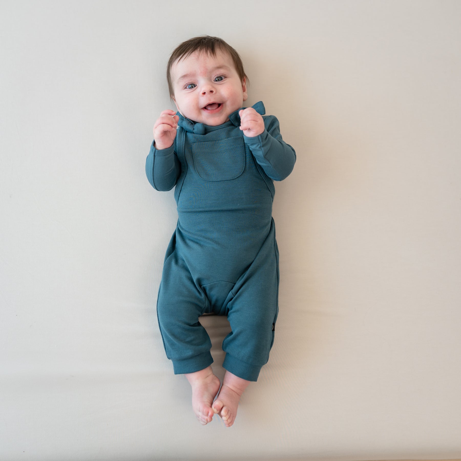 Infant laying down on a Bisque blanket wearing the Bamboo Jersey Overall in Atlantic and matching long sleeve bodysuit