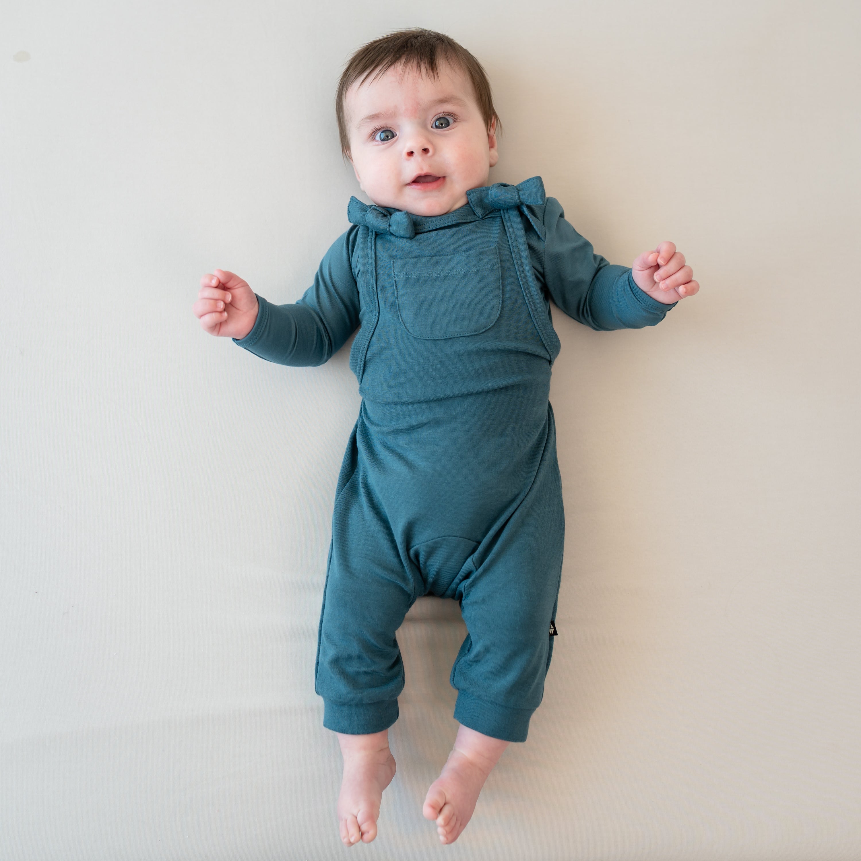 Infant laying on a Bisque Blanket wearing the Bamboo Jersey Overall in Atlantic with a matching long sleeve bodysuit on underneath