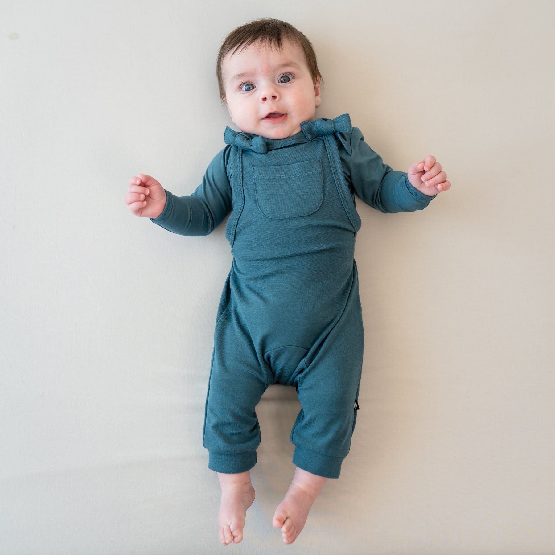 Infant laying on a Bisque Blanket wearing the Bamboo Jersey Overall in Atlantic with a matching long sleeve bodysuit on underneath