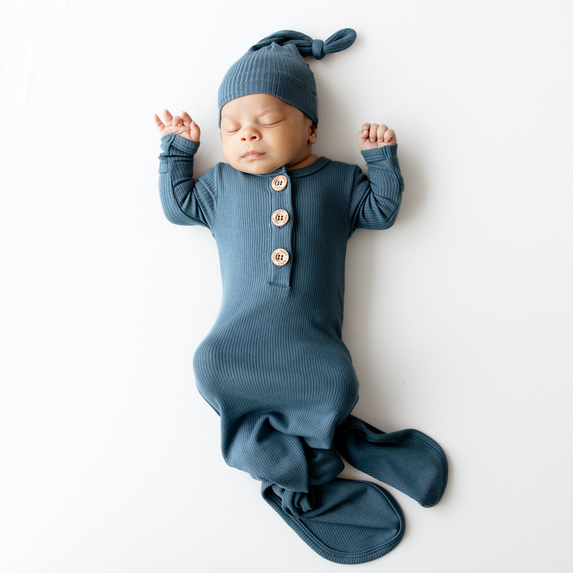 Sleeping infant wearing the Ribbed Knotted Gown with Hat Set in Atlantic laying on a light colored background