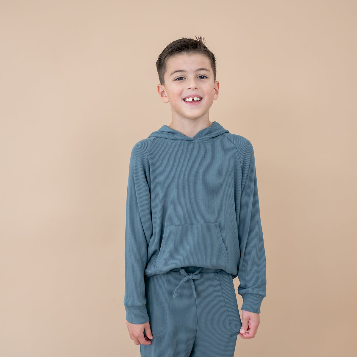 Smiling boy wearing the Ribbed Hoodie in Atlantic paired with matching ribbed jogger pant in front of a taupe background