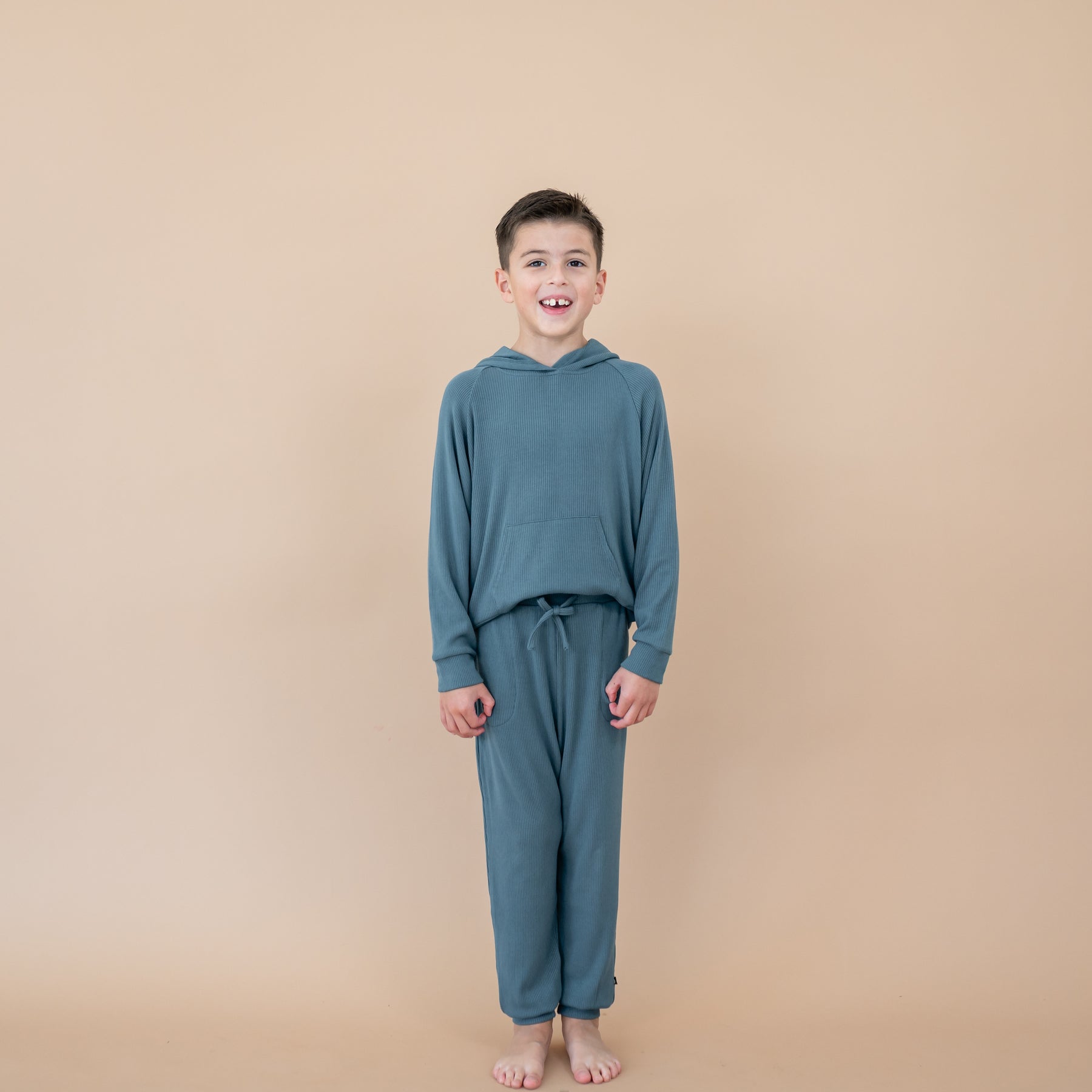 Smiling boy model wearing the Ribbed Jogger Pant in Atlantic paired with the matching ribbed hoodie in front of a taupe background