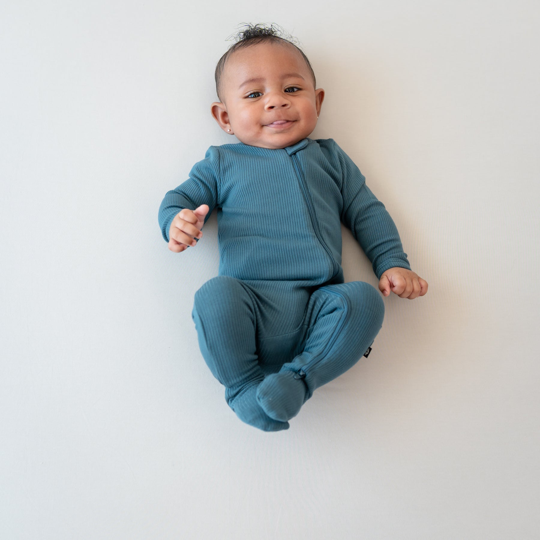 Smiling infant sticking their tongue out wearing the Ribbed Zipper Footie in Atlantic on a bisque blanket