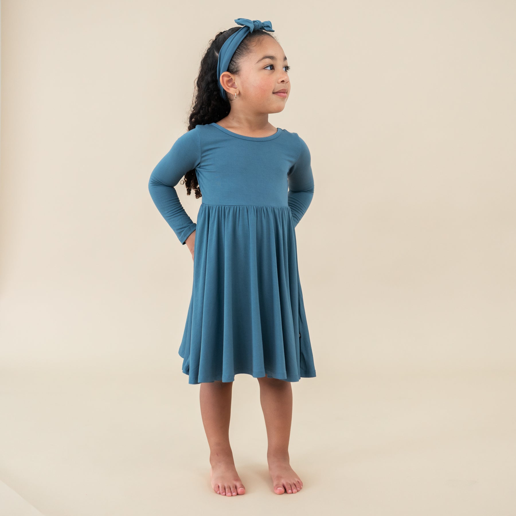 Girl standing with hands on hips looking off to the side wearing the Long Sleeve Twirl Dress in Atlantic with matching knotted bow headband