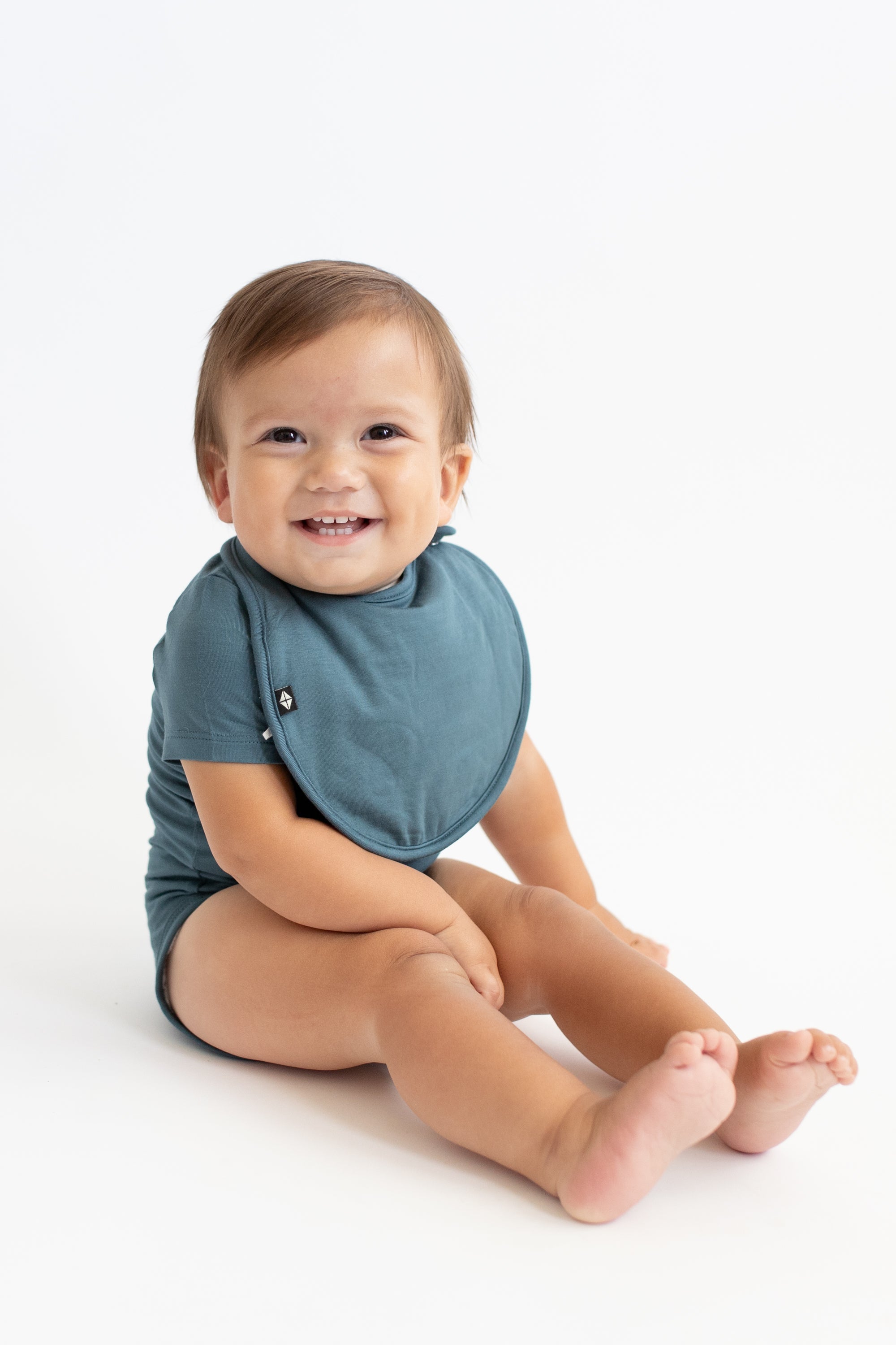 Smiling infant sitting with his legs out in front of him wearing the Cushy Bib in Atlantic with matching short sleeve bodysuit