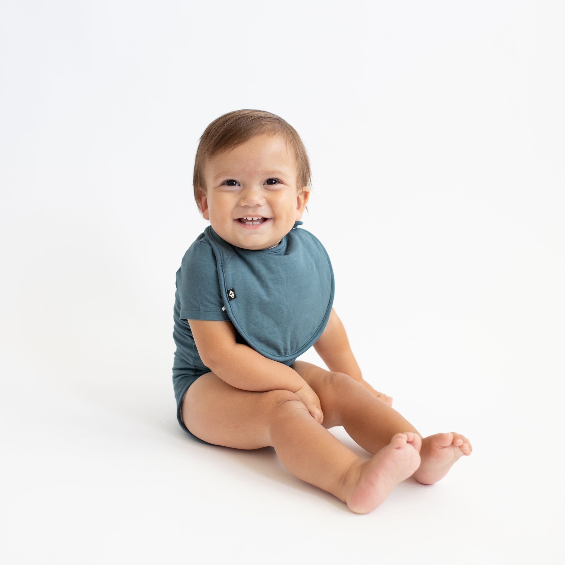 Smiling infant sitting with his legs out in front of him wearing the Cushy Bib in Atlantic with matching short sleeve bodysuit