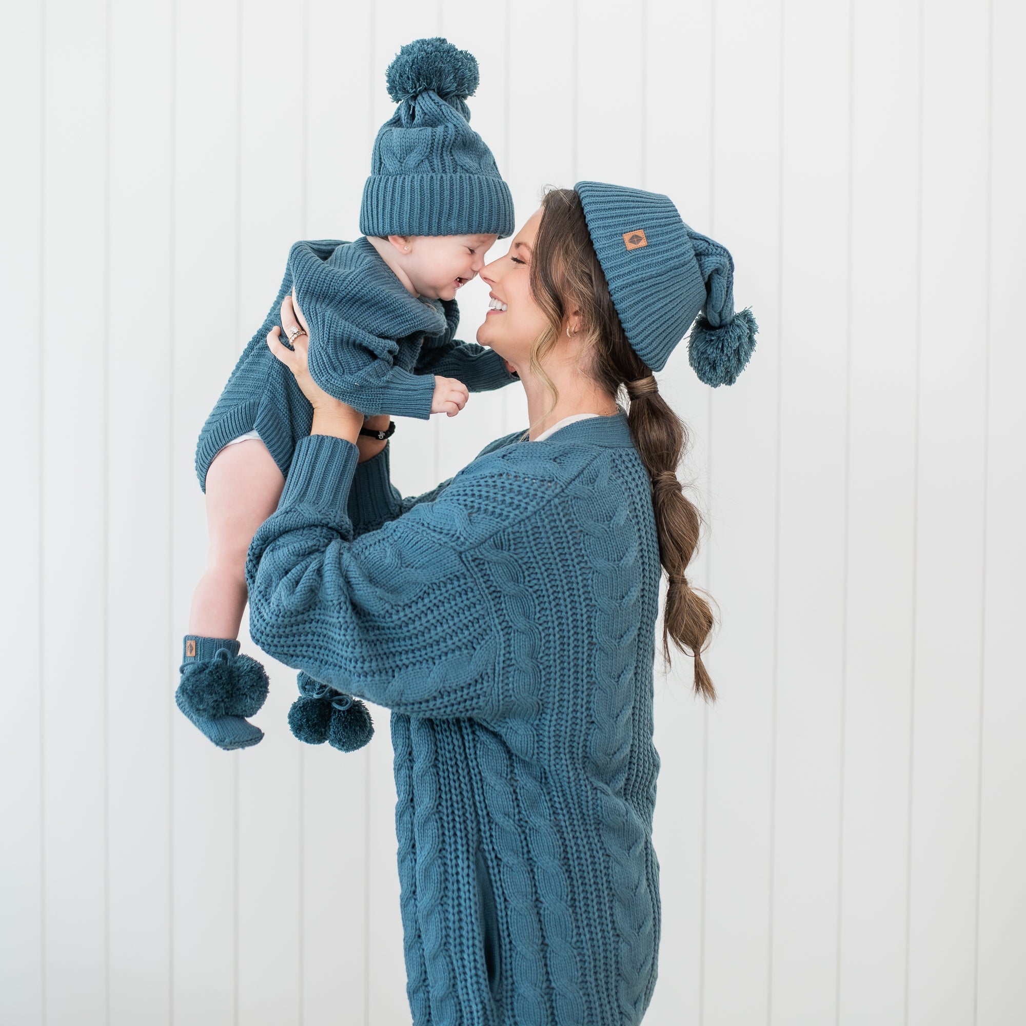 Mother holding child to her face touching noses. The mother wearing the Cable Knit Adult Pom Beanie in Atlantic with matching cable knit cardigan while the child is wearing a matching Cable Knit Pom Beanie in Atlantic, Chunky knit romper and knit booties