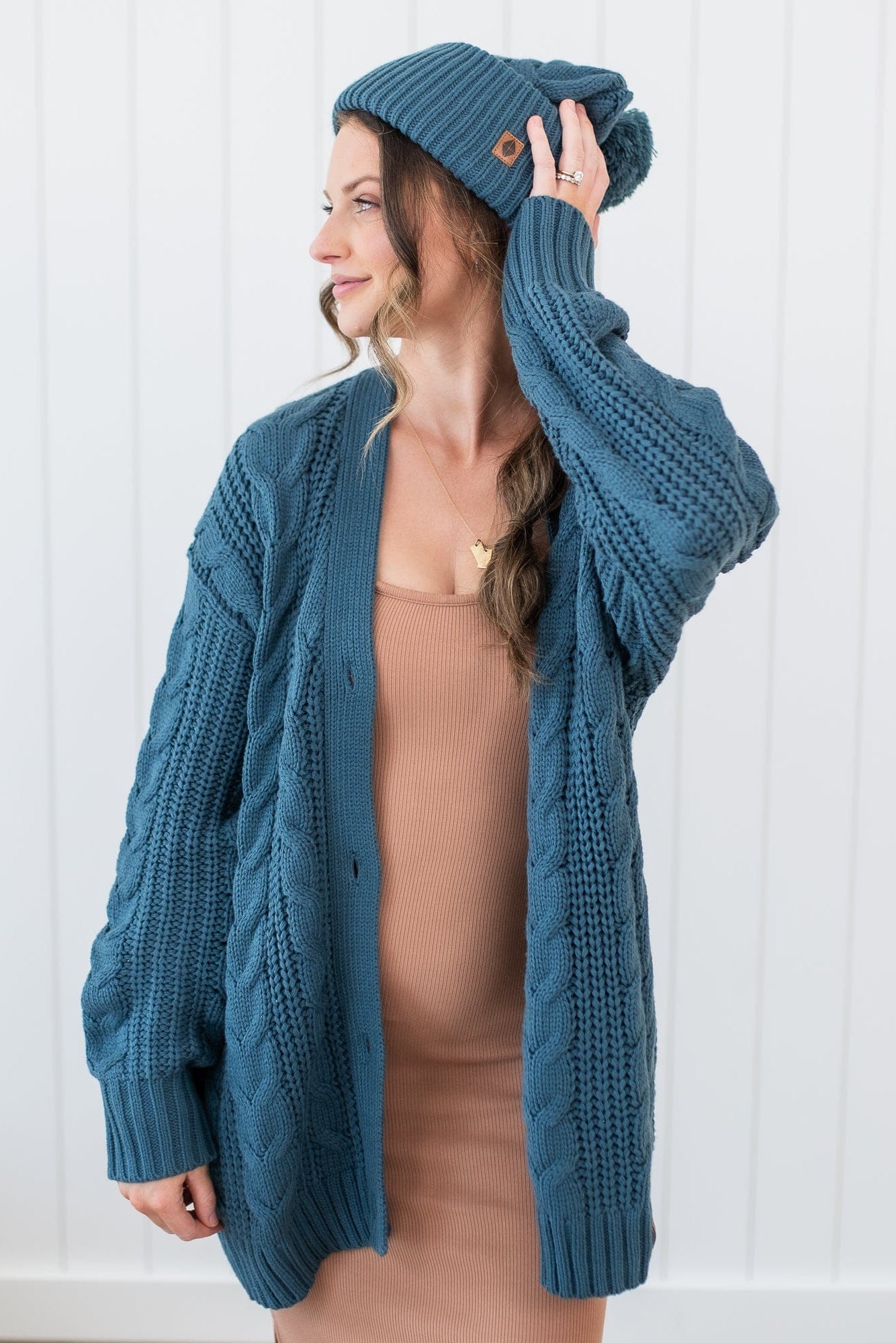 Female model wearing the Cable Knit Adult Cardigan in Atlantic paired with a matching chunky knit pom beanie and latte cami dress