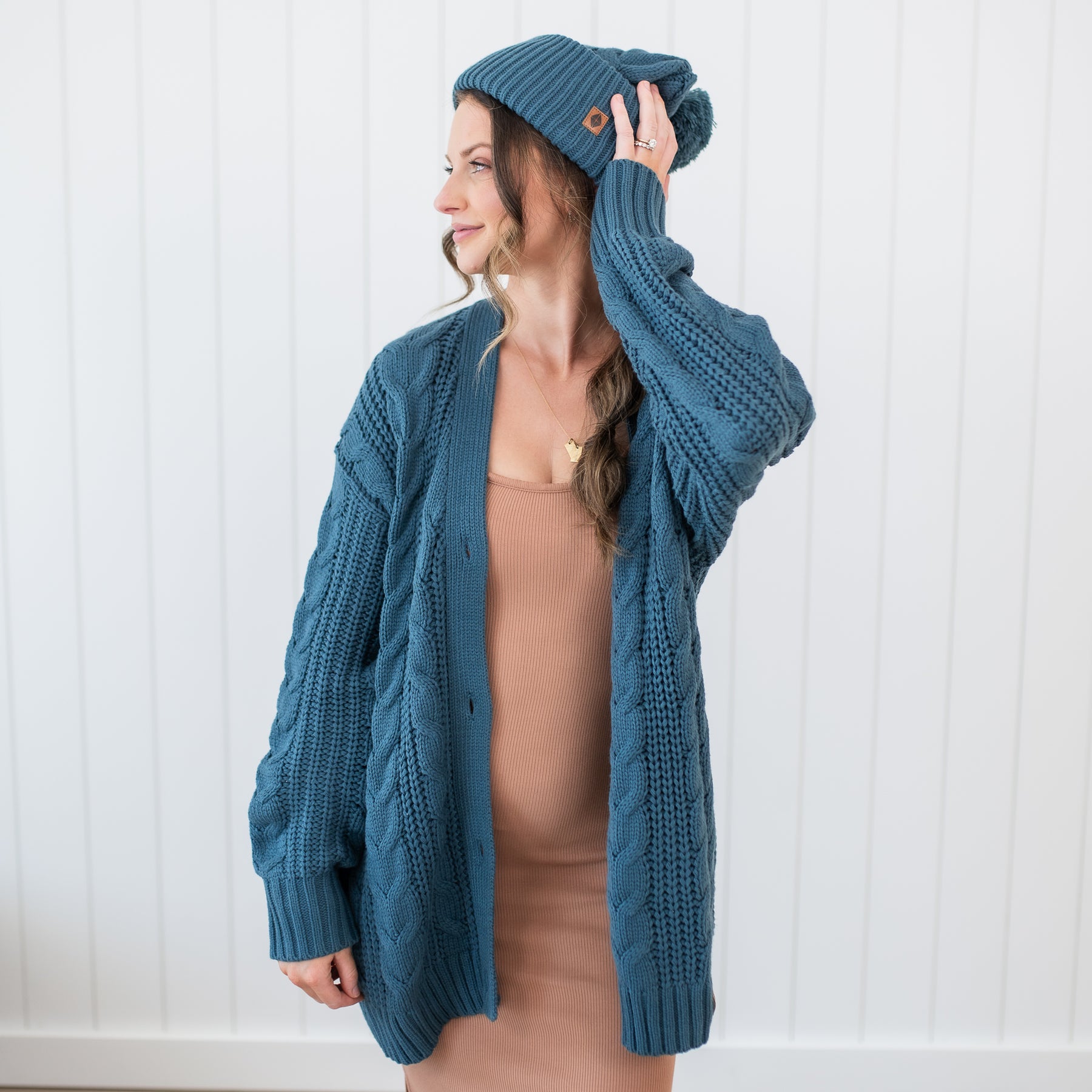Female model wearing the Cable Knit Adult Cardigan in Atlantic paired with a matching chunky knit pom beanie and latte cami dress