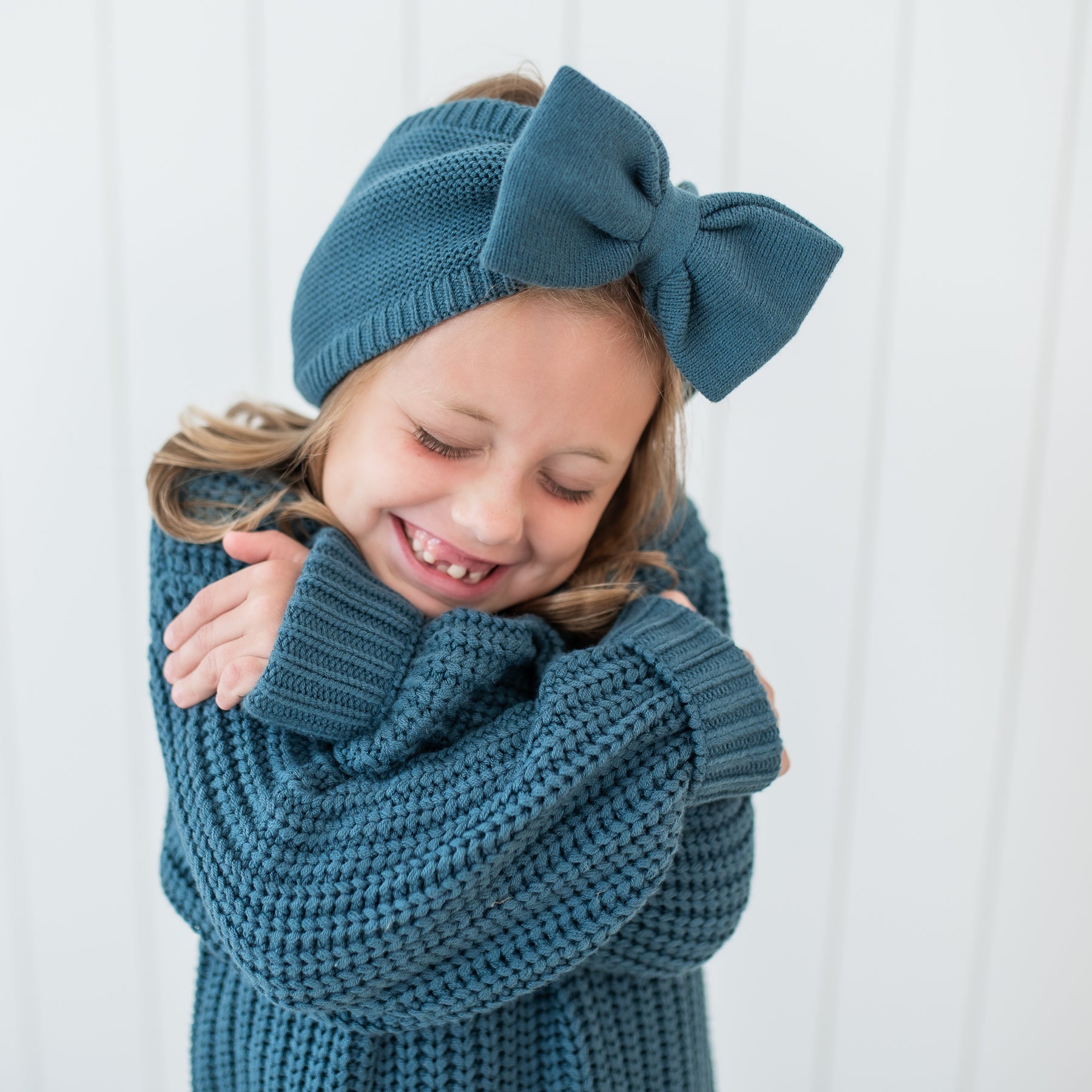 Young girl hugging herself wearing the Chunky Knit Bow Headband in Atlantic and matching Chunky Knit Sweater