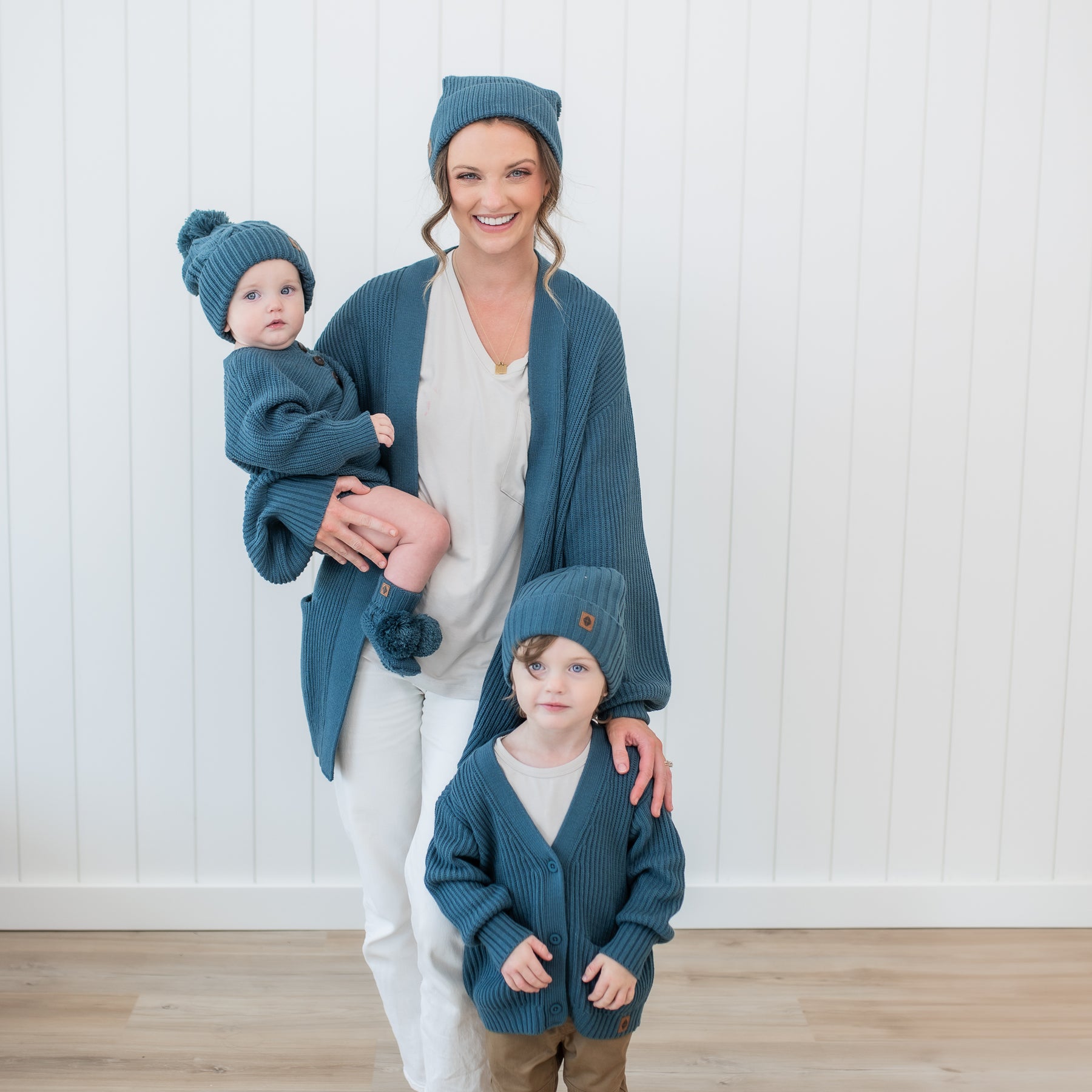 Family matching in Atlantic Chunky knit items. Toddler wearing the Knit Booties in Atlantic, chunky knit romper and cable knit pom beanie being held by mother who is wearing a chunky knit pom beanie and chunky knit women's oversized cardigan. Older child is standing beside mother wearing the chunky knit beanie and chunky knit oversized cardigan