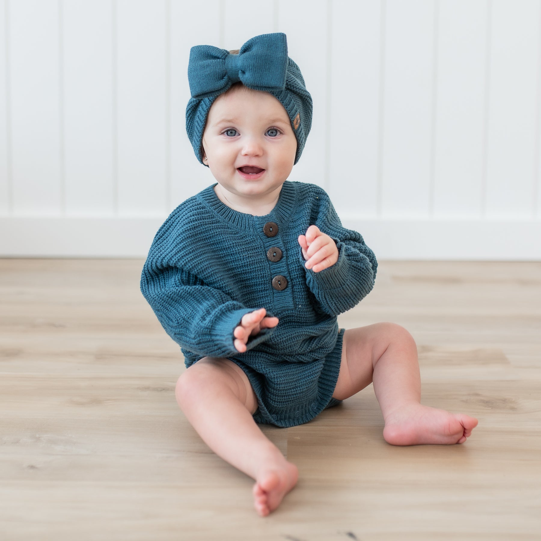 Toddler sitting on the floor wearing the Chunky Knit Romper in Atlantic paired with a Chunky knit bow headband