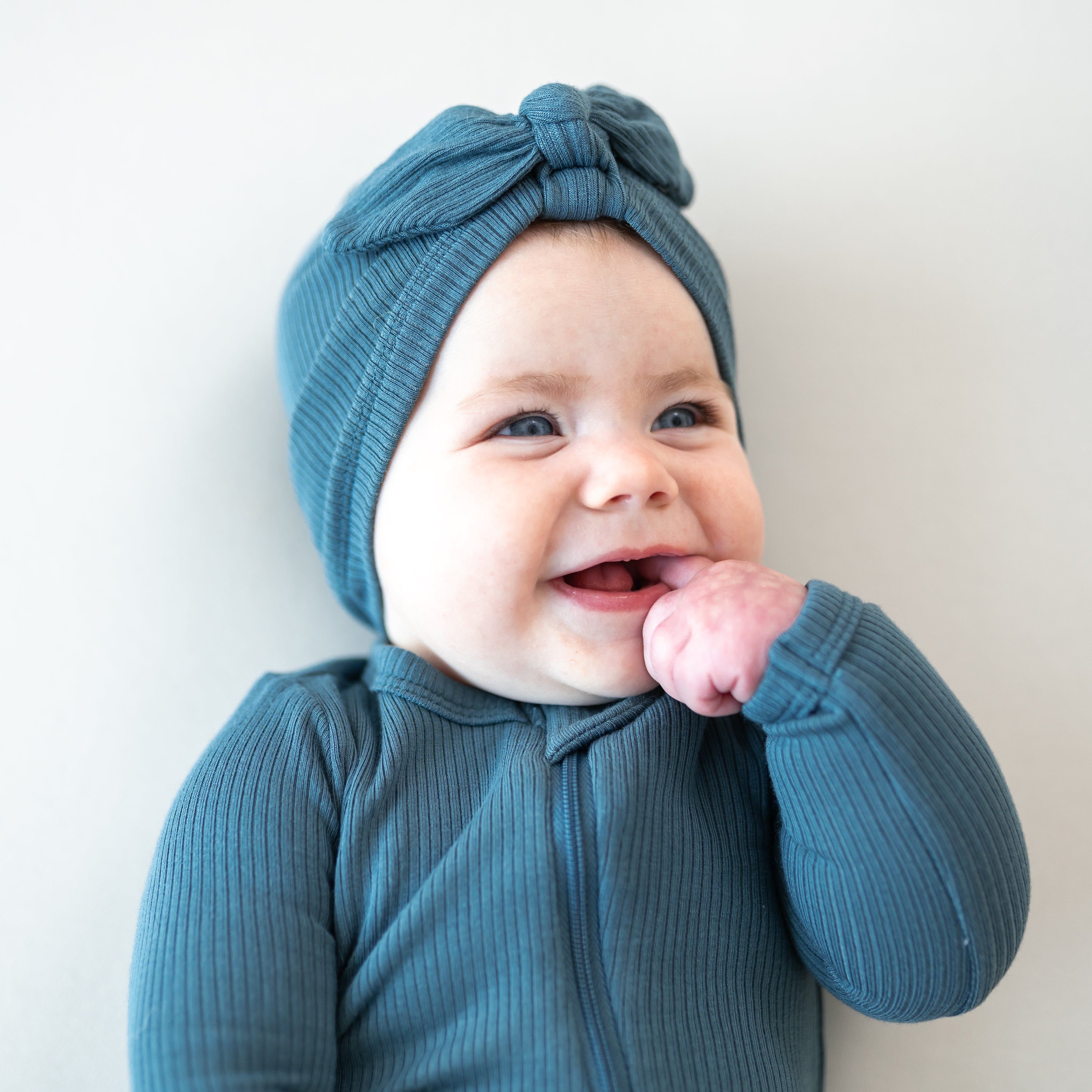 Smiling infant with one finger in her mouth wearing the Ribbed Headwrap in Atlantic and matching ribbed zippered romper