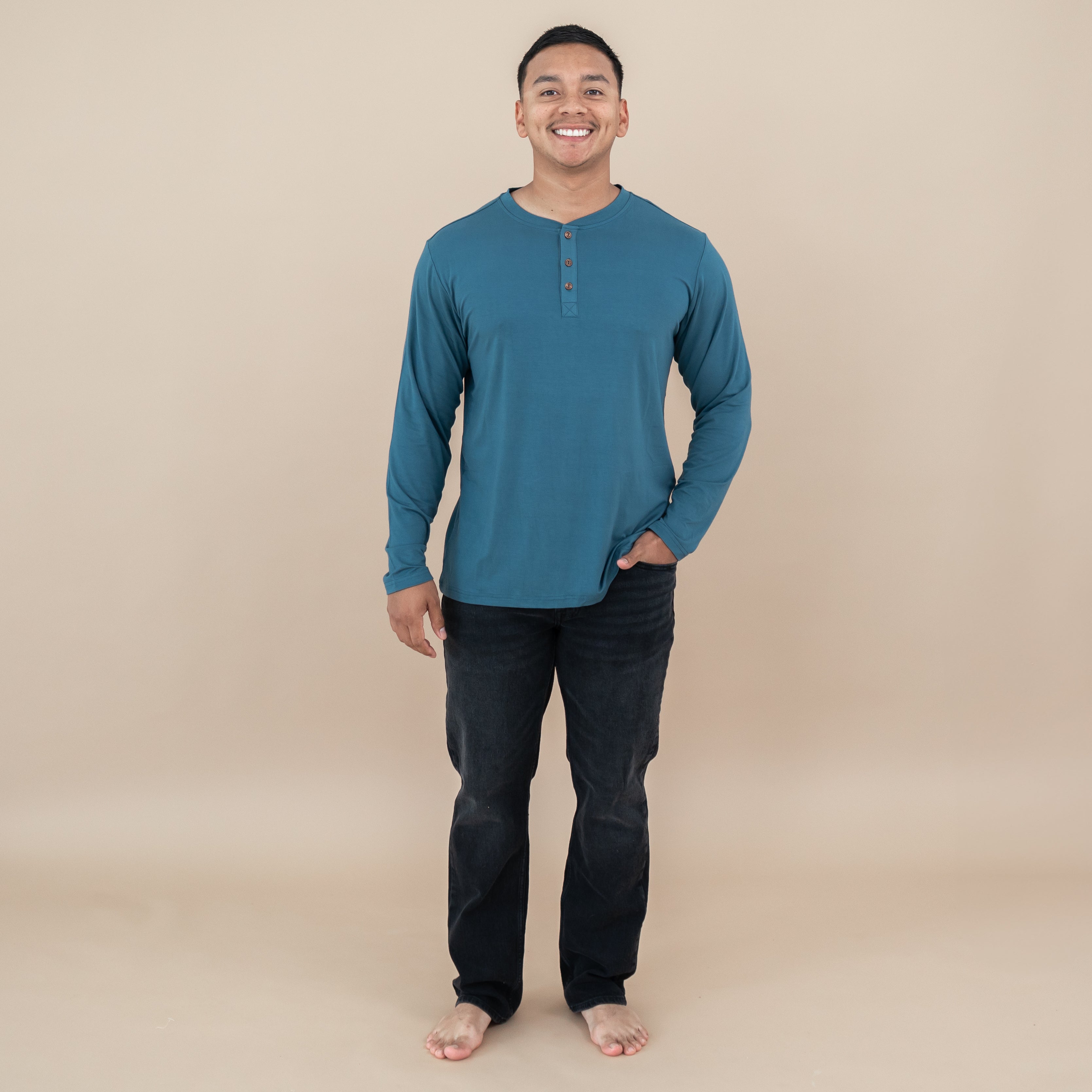 Smiling male model standing wearing the Men's Long Sleeve Henley Top in Atlantic with one hand in his black jeans pocket