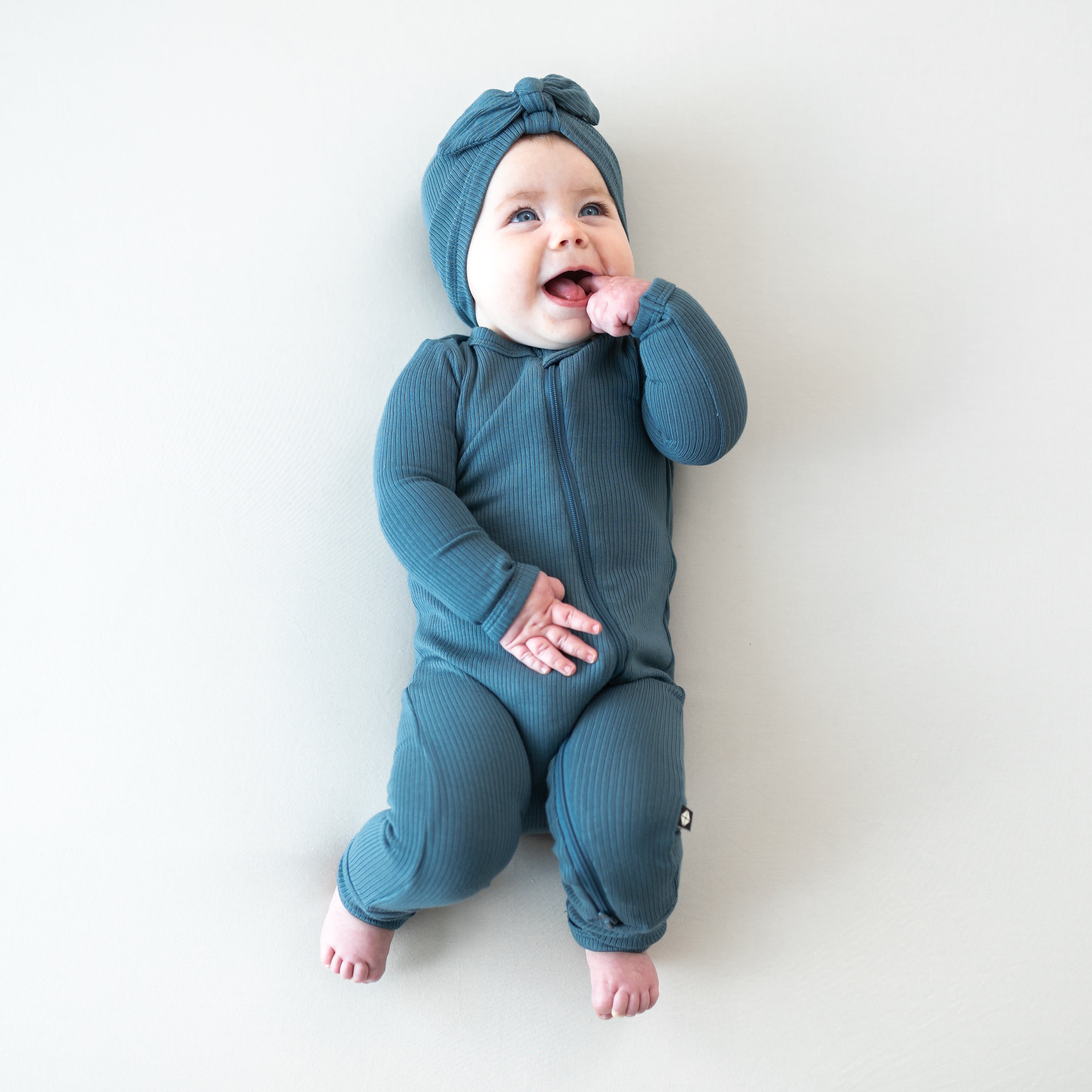 Smiling infant laying down on a light background wearing the Ribbed Headwrap in Atlantic paired with a matching ribbed zipper romper