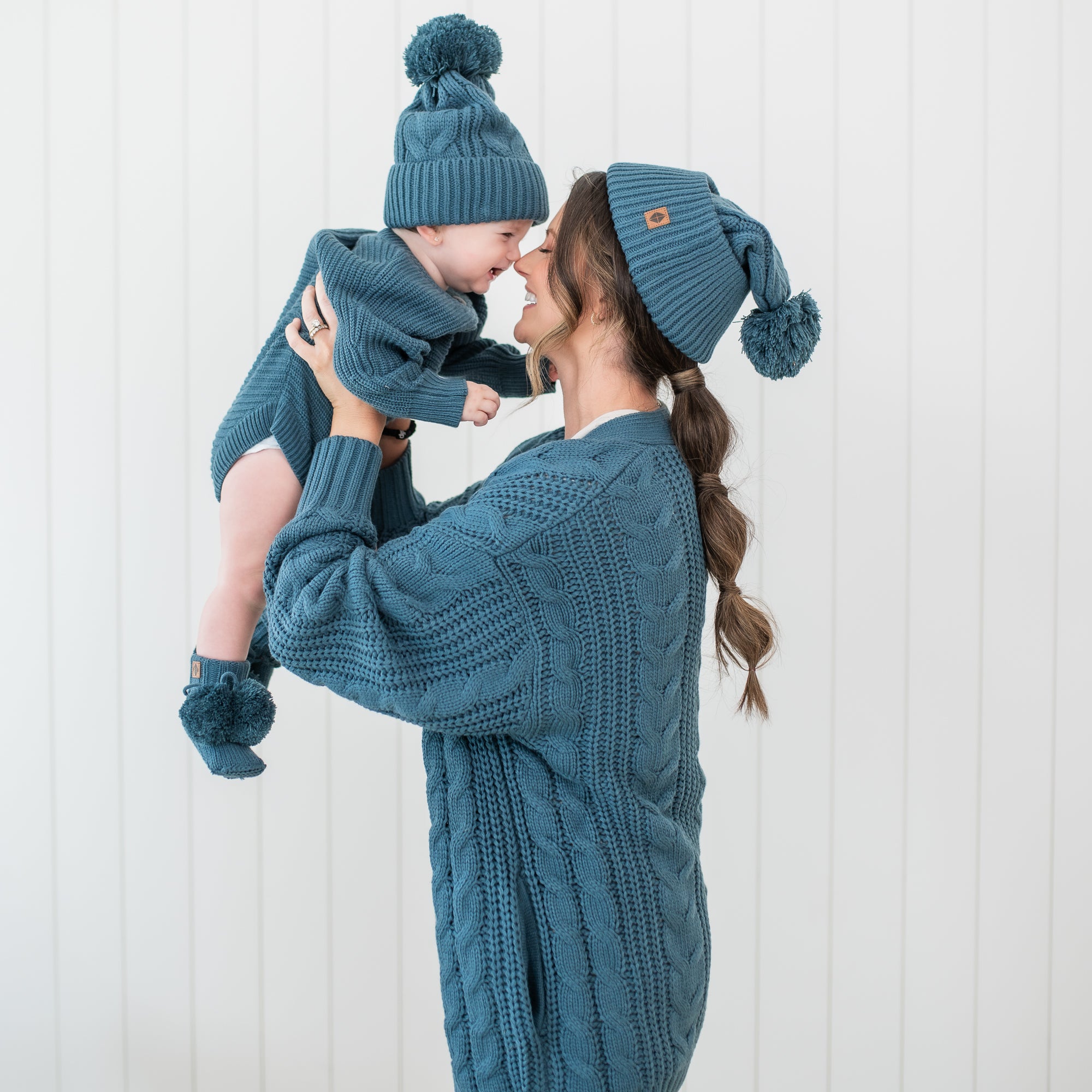 Female model holding her son up to her face with their noses touching. Mother wearing the Cable Knit Adult Cardigan in Atlantic and Cable Knit Pom beanie with her child matching in an Atlantic chunky knit romper, knit booties and cable knit pom beanie